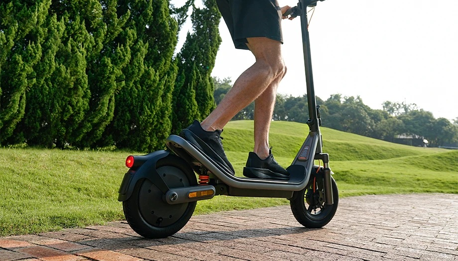 Mężczyzna jedzie na hulajnodze elektrycznej po wybrukowanej ścieżce, z zielonymi drzewami i wzgórzami w tle.