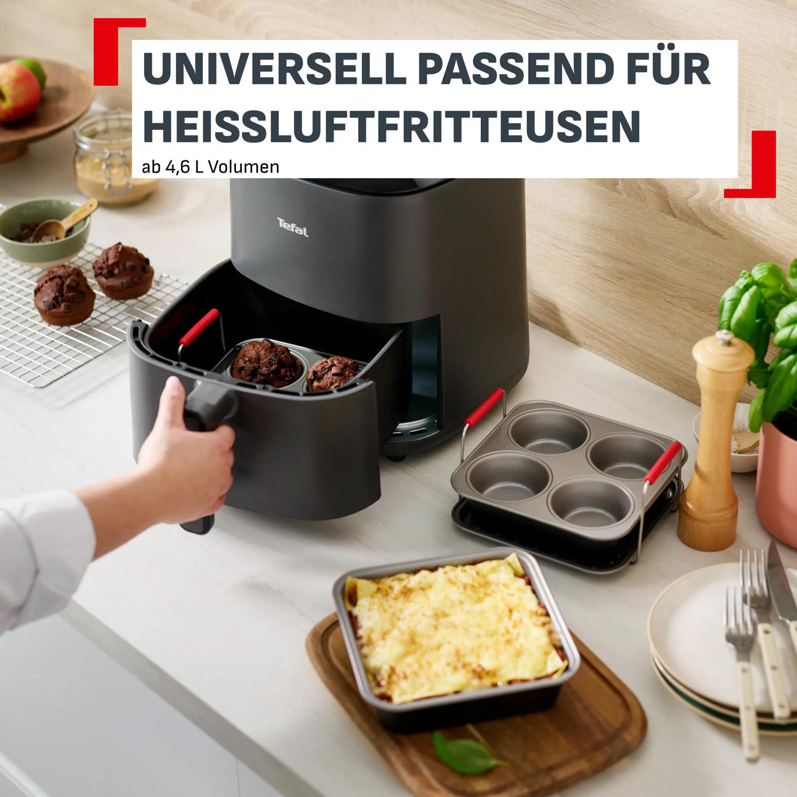 Eine Hand zieht einen Heißluftfritteusenkorb mit Muffins heraus. Daneben sind weitere Backutensilien.