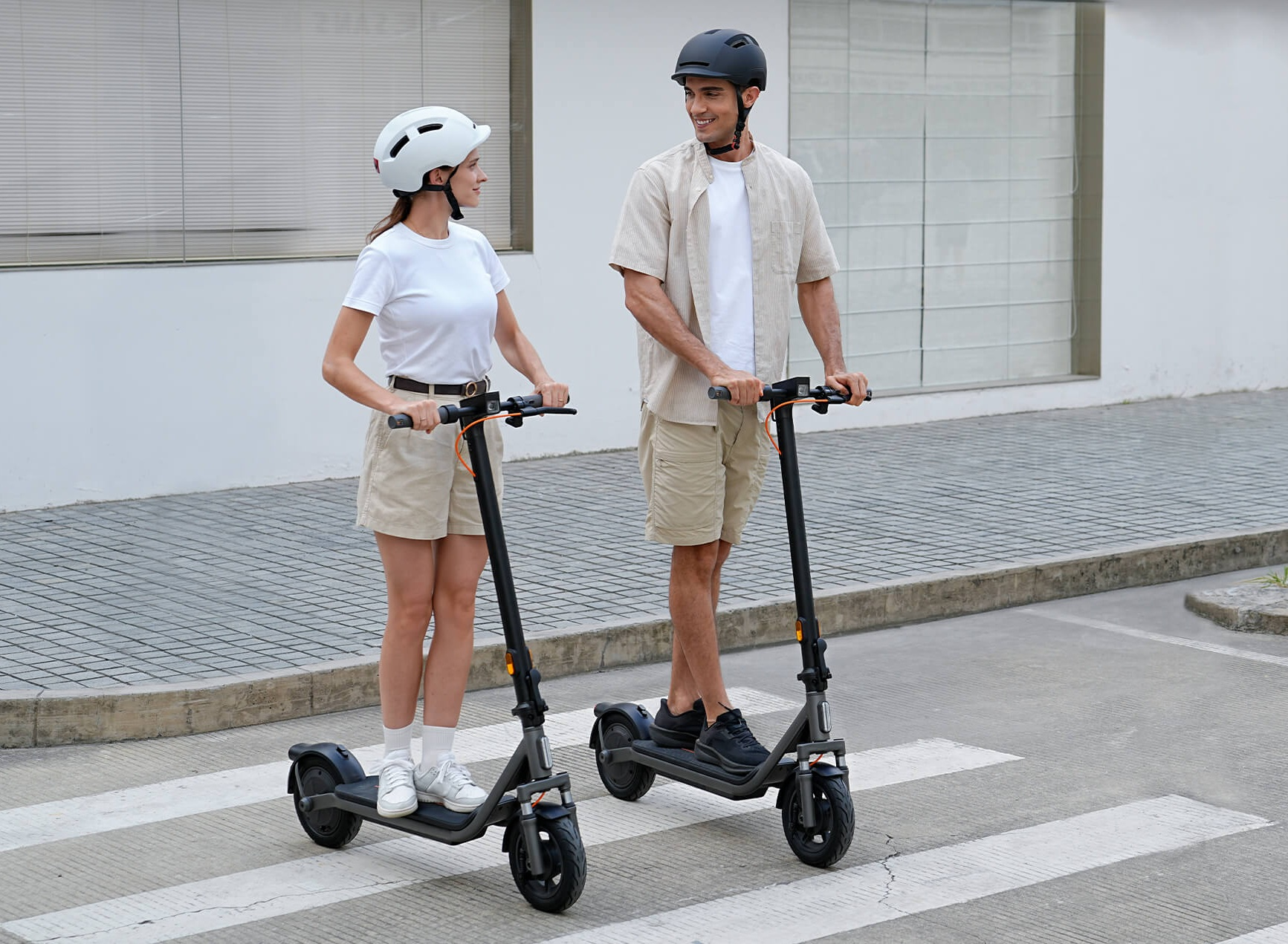 Mężczyzna i kobieta w kaskach na hulajnogach elektrycznych na przejściu dla pieszych.