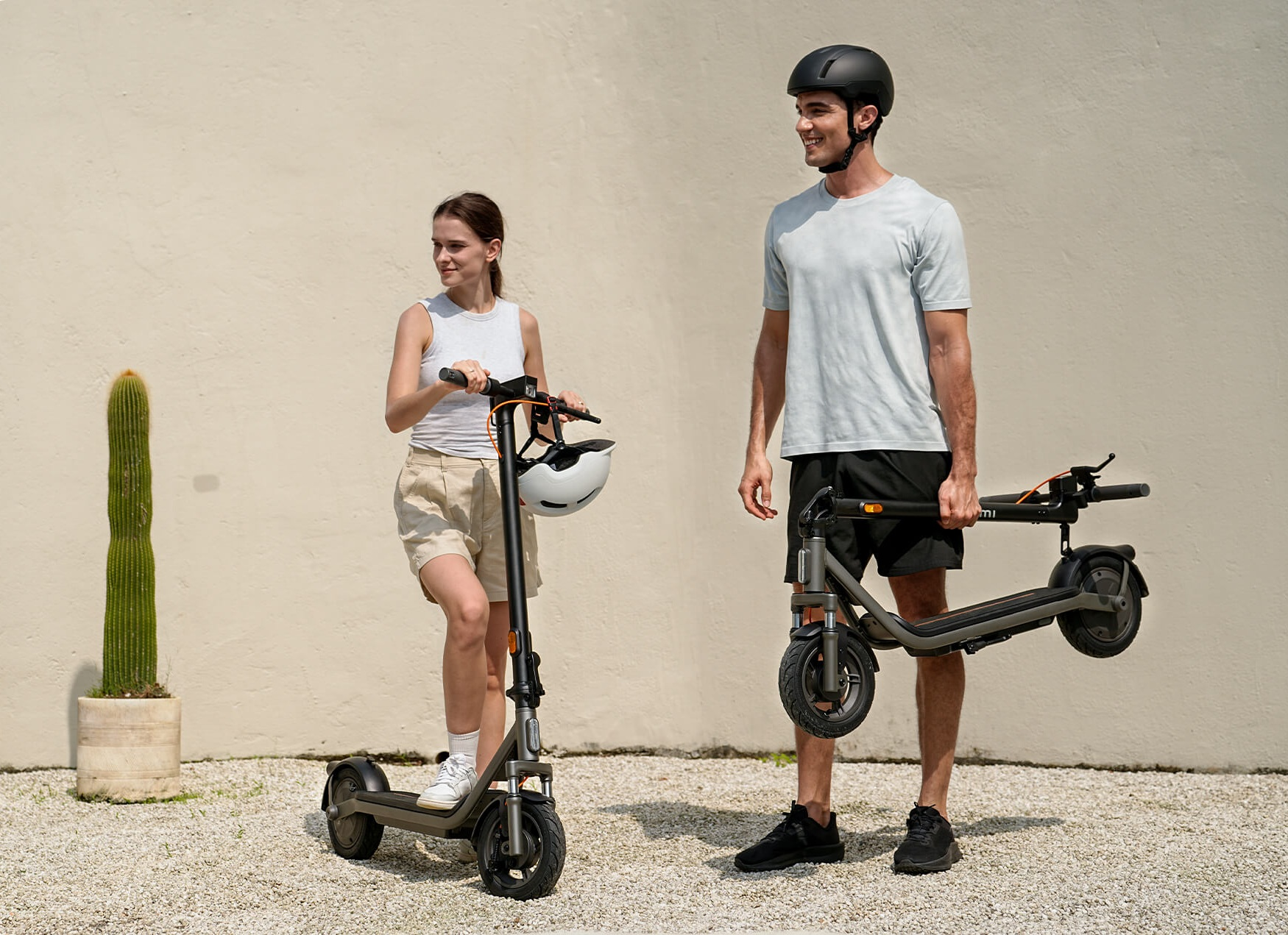 Mężczyzna i kobieta z hulajnogami elektrycznymi przed ścianą, mężczyzna trzyma złożoną hulajnogę.