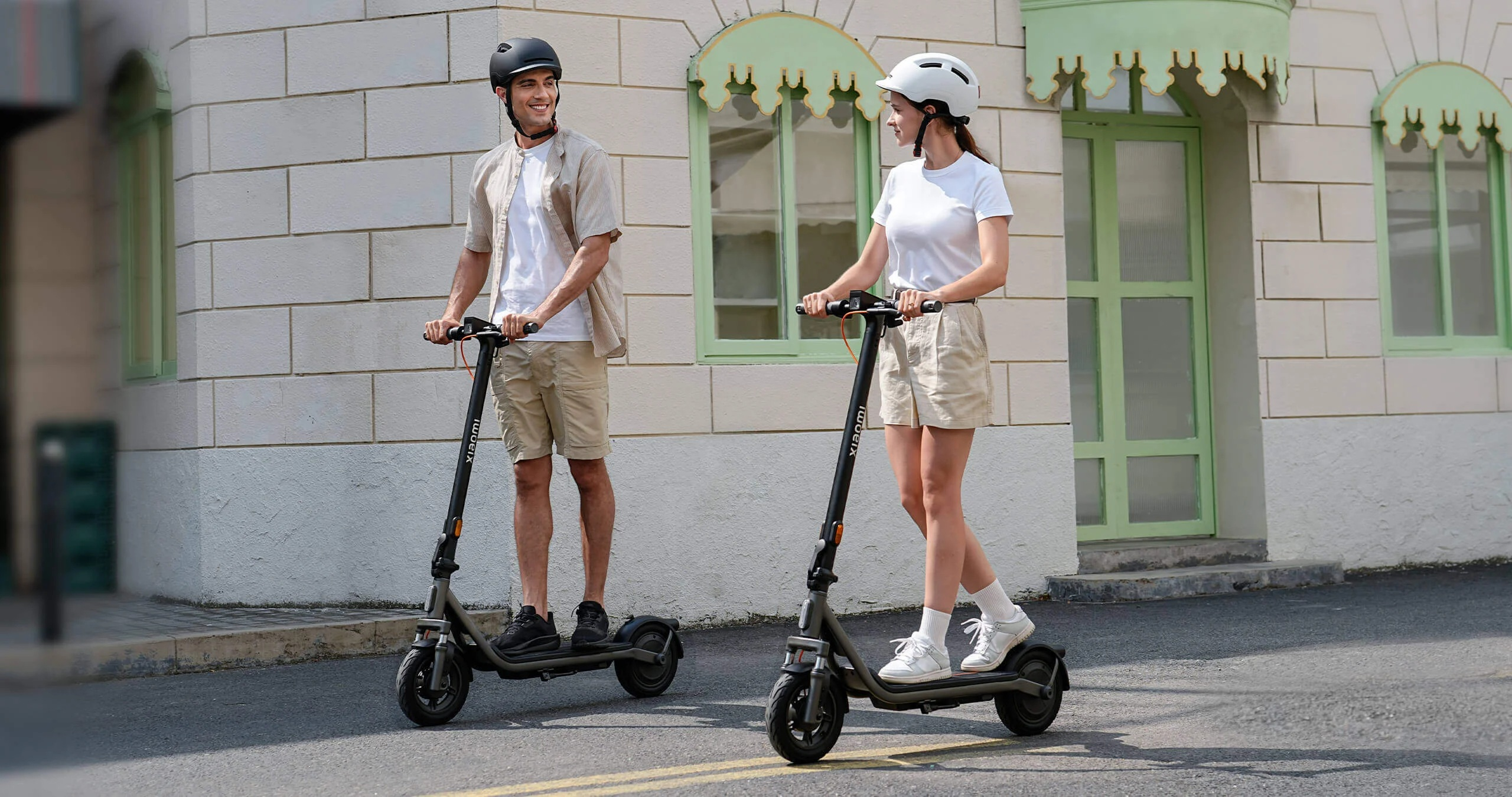 Mężczyzna i kobieta na hulajnogach elektrycznych przed budynkiem z zielonymi oknami.