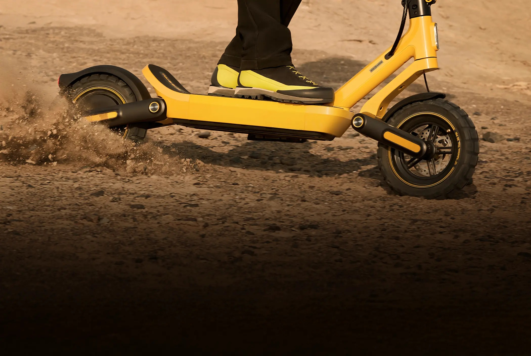 Żółty skuter elektryczny jedzie po nierównym terenie, wzbijając kurz.