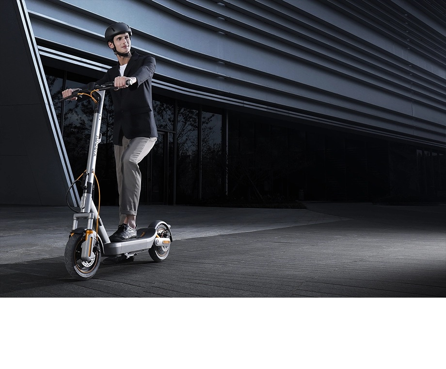 Mężczyzna w kasku i garniturze jedzie srebrną hulajnogą elektryczną po chodniku.