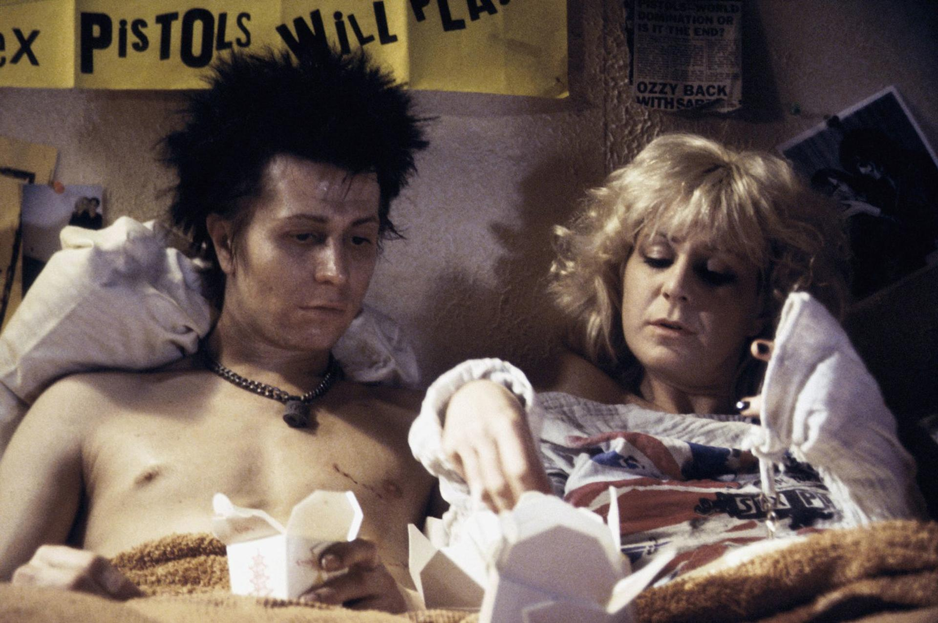 Ein Mann und eine Frau liegen im Bett, umgeben von Punk-Devotionalien und Essensbehältern.