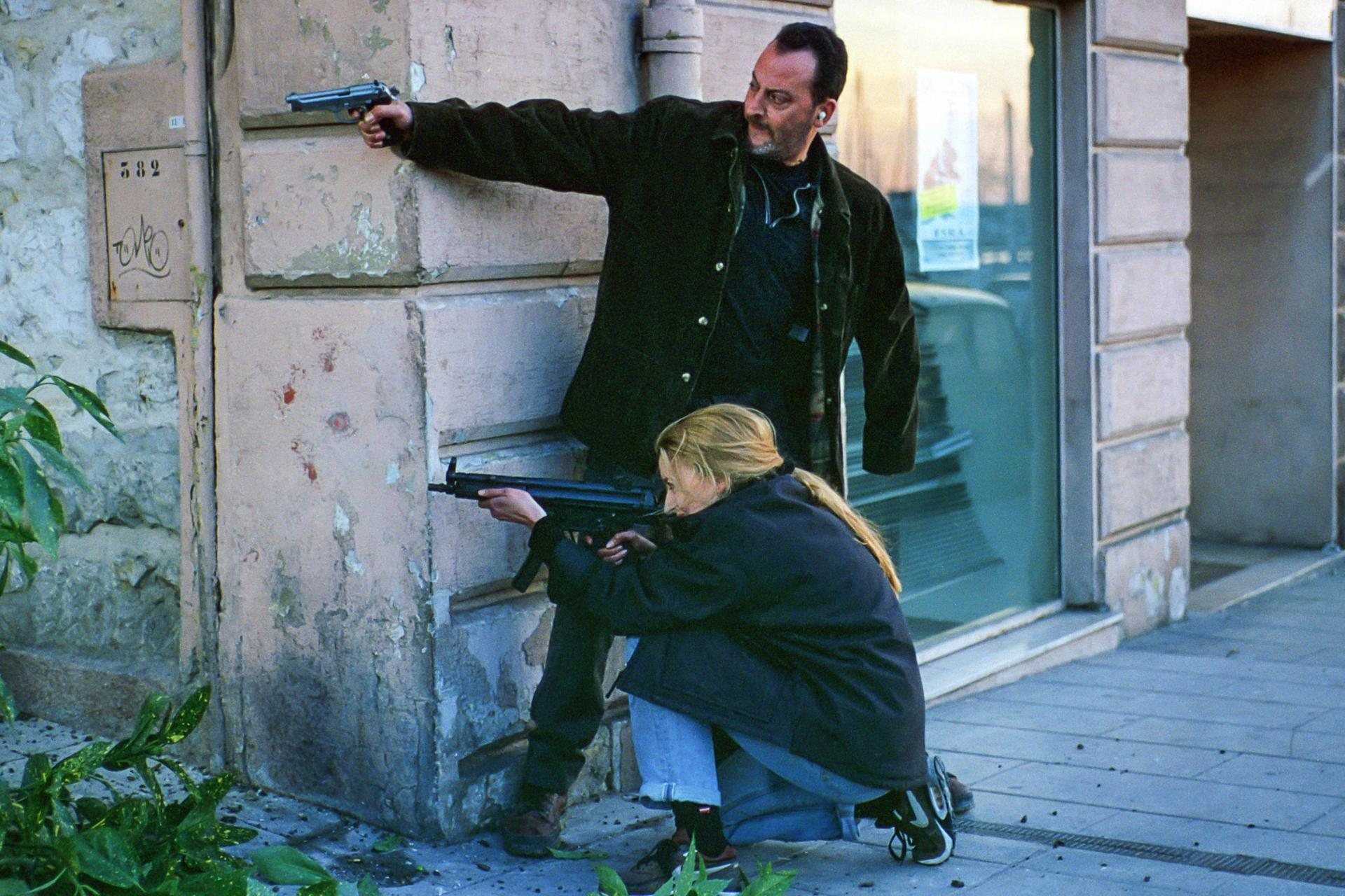 Ein Mann mit Waffe lehnt an einer Wand, eine Frau kniet mit Waffe.