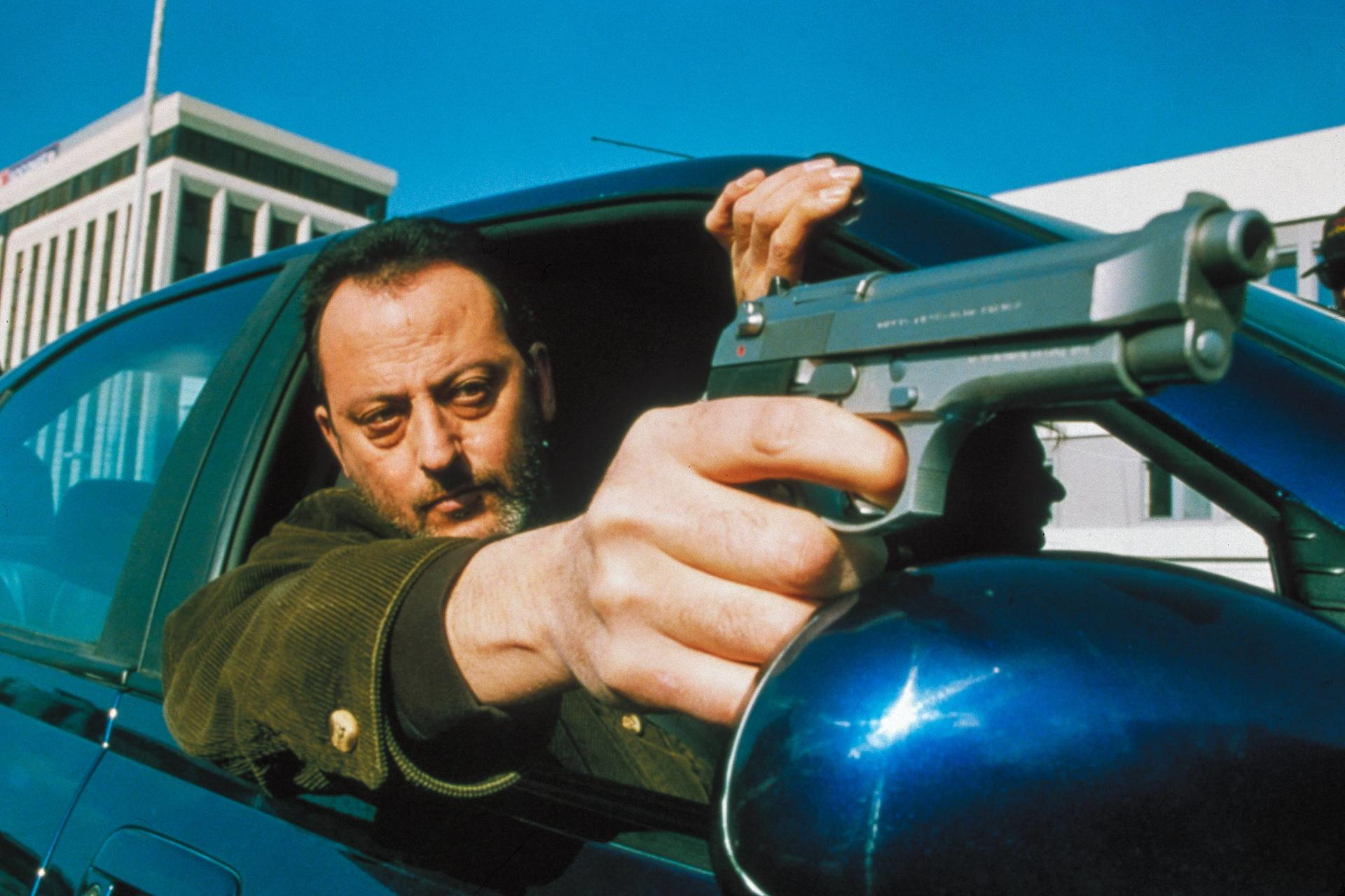 Mann im Auto hält eine Waffe, Blick nach vorne gerichtet.