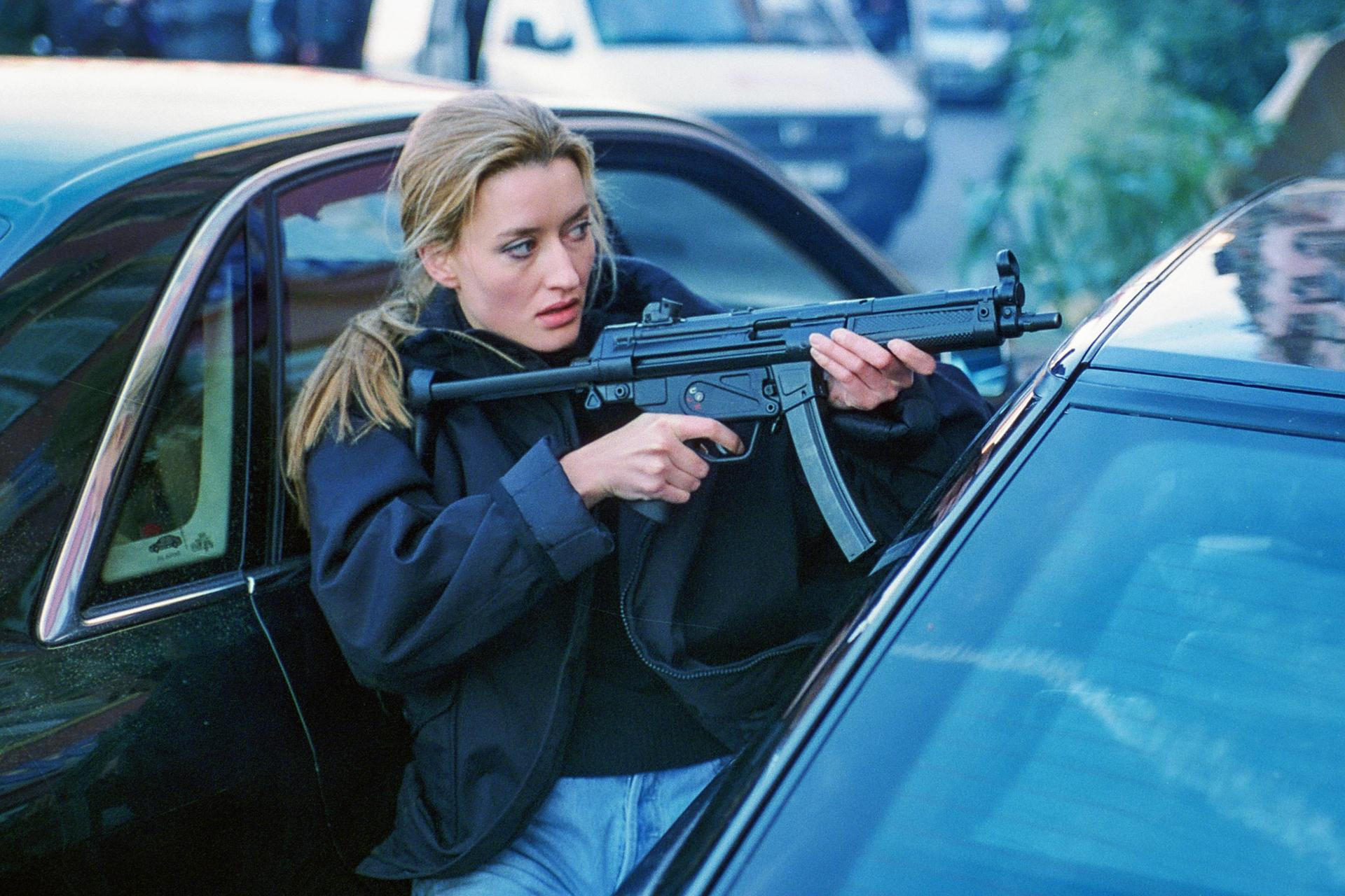Eine Frau mit blonden Haaren hält eine Maschinenpistole, während sie aus einem Auto lugt.