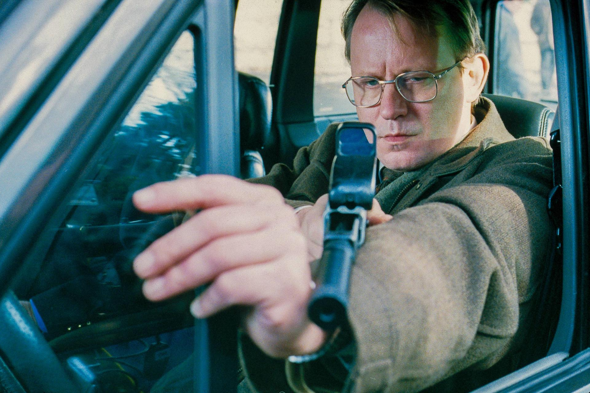 Mann mit Brille zielt mit Waffe aus Autofenster.