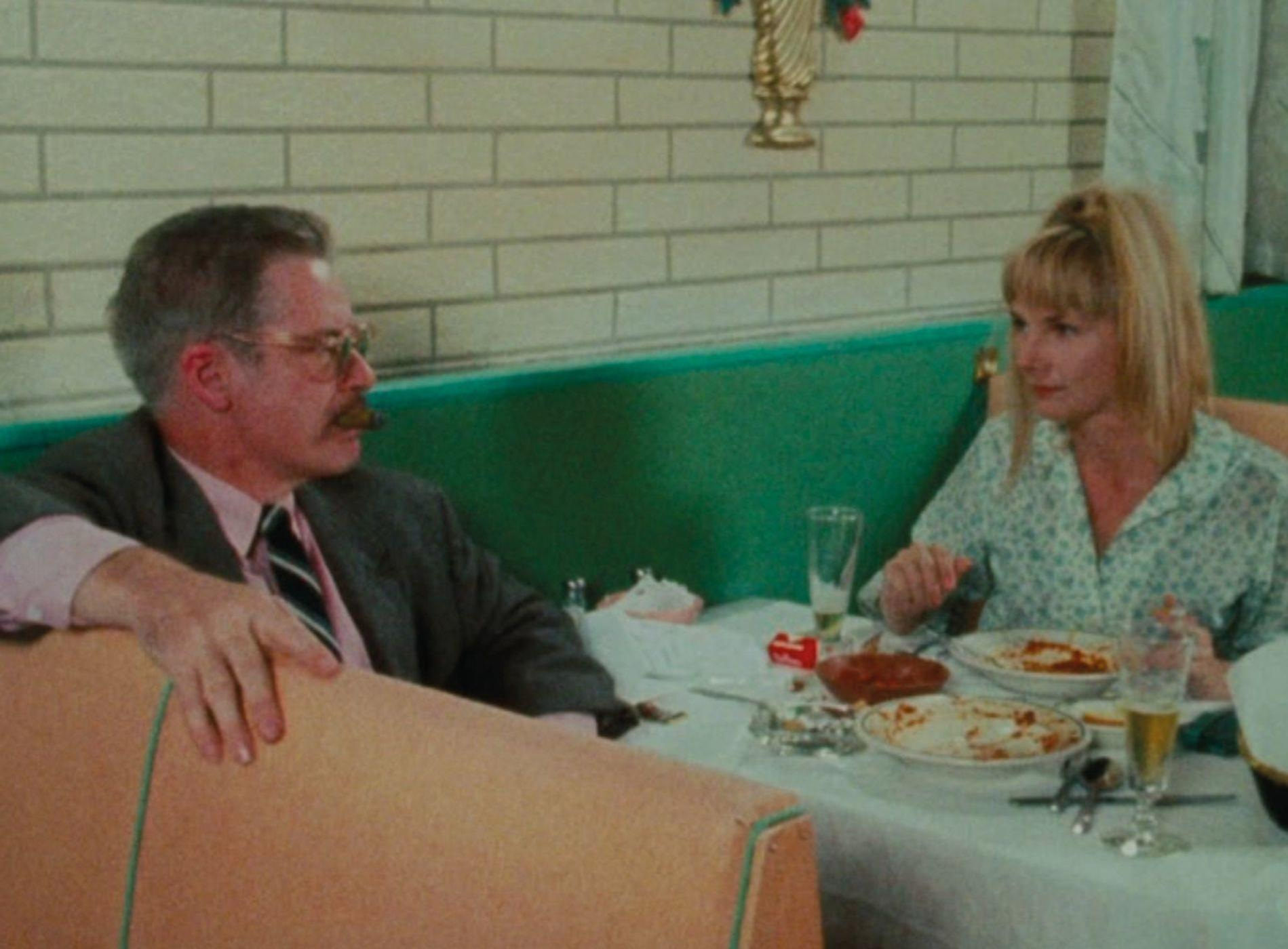 Mann und Frau essen an einem Tisch in einer Nische. Mann hat Schnurrbart, Brille, Anzug. Frau hat blonde Haare.