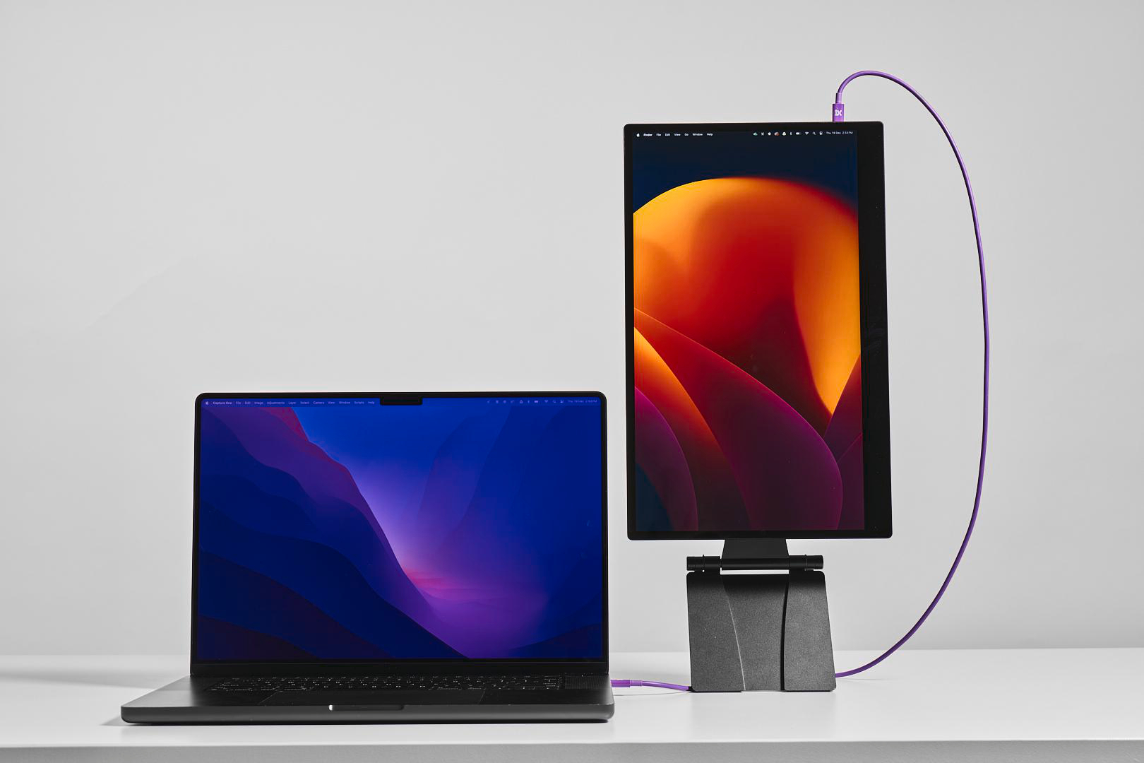 Ein Laptop und ein vertikaler Monitor zeigen bunte abstrakte Hintergrundbilder. Ein lila Kabel verbindet sie.