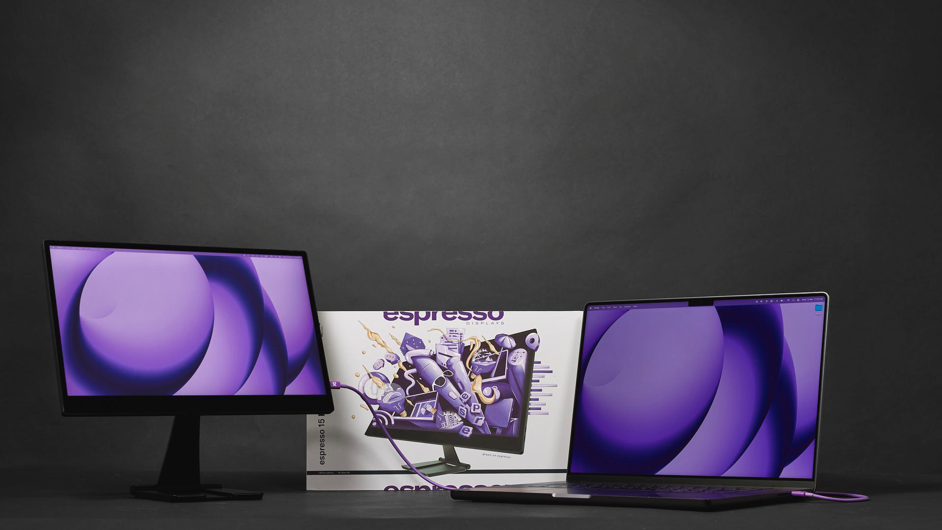 Ein Laptop und zwei Monitore mit violetten Hintergründen stehen auf einem dunklen Tisch.
