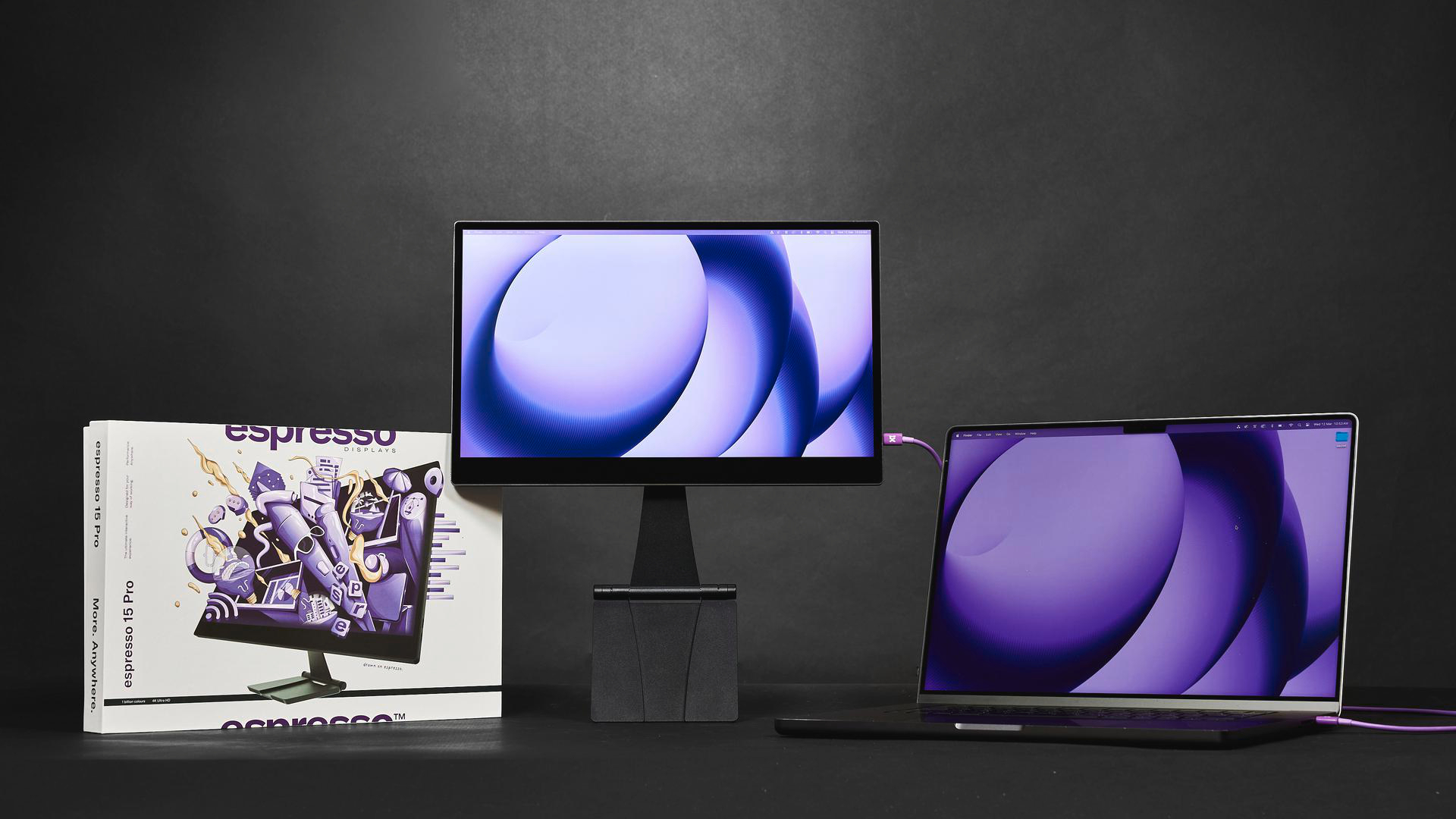 Ein Laptop, ein Monitor und eine Verpackung mit lila abstrakter Kunst auf dunklem Hintergrund.
