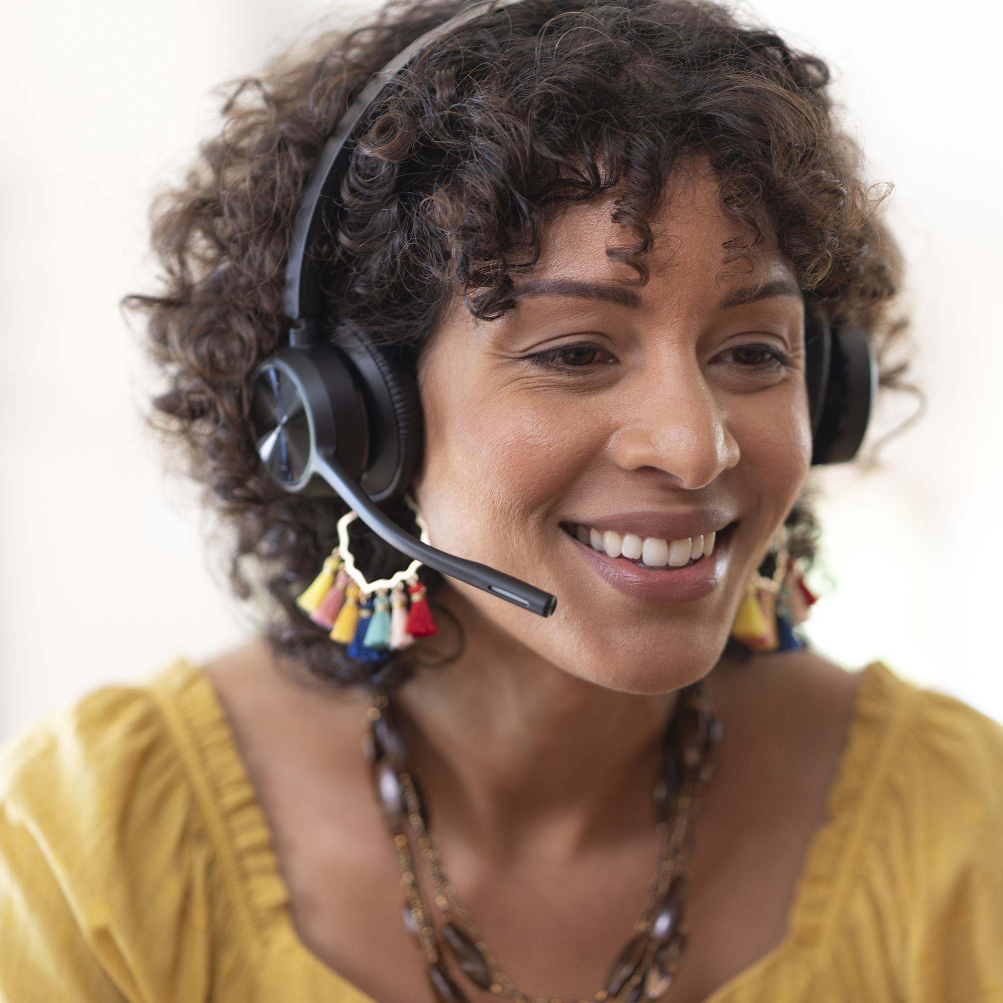 Eine lächelnde Frau mit lockigem Haar trägt ein Headset und ein gelbes Oberteil.