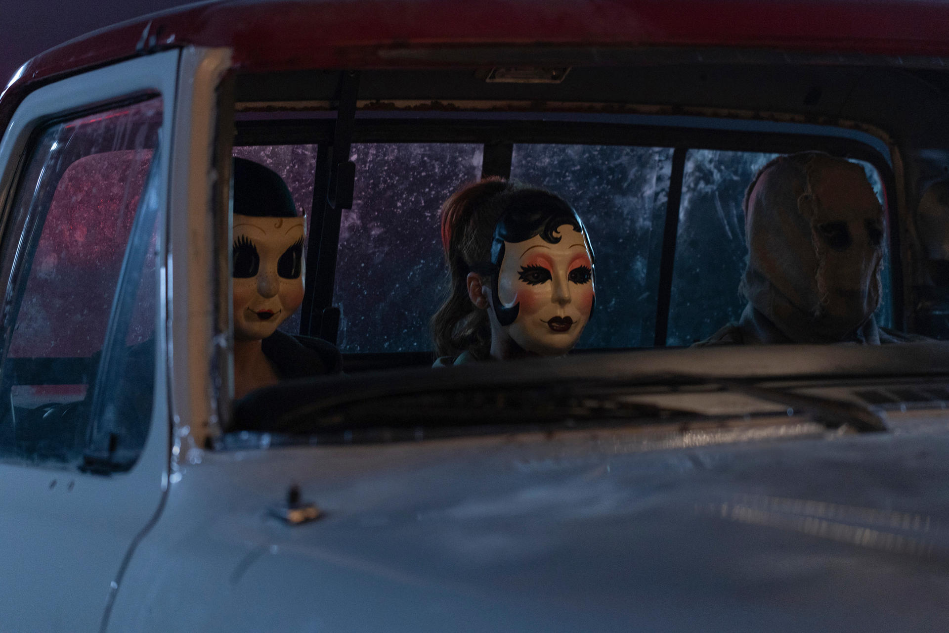Drei Personen in Masken sitzen in einem Auto. Links eine Puppenmaske, Mitte eine Theatermaske, rechts eine Sackmaske.