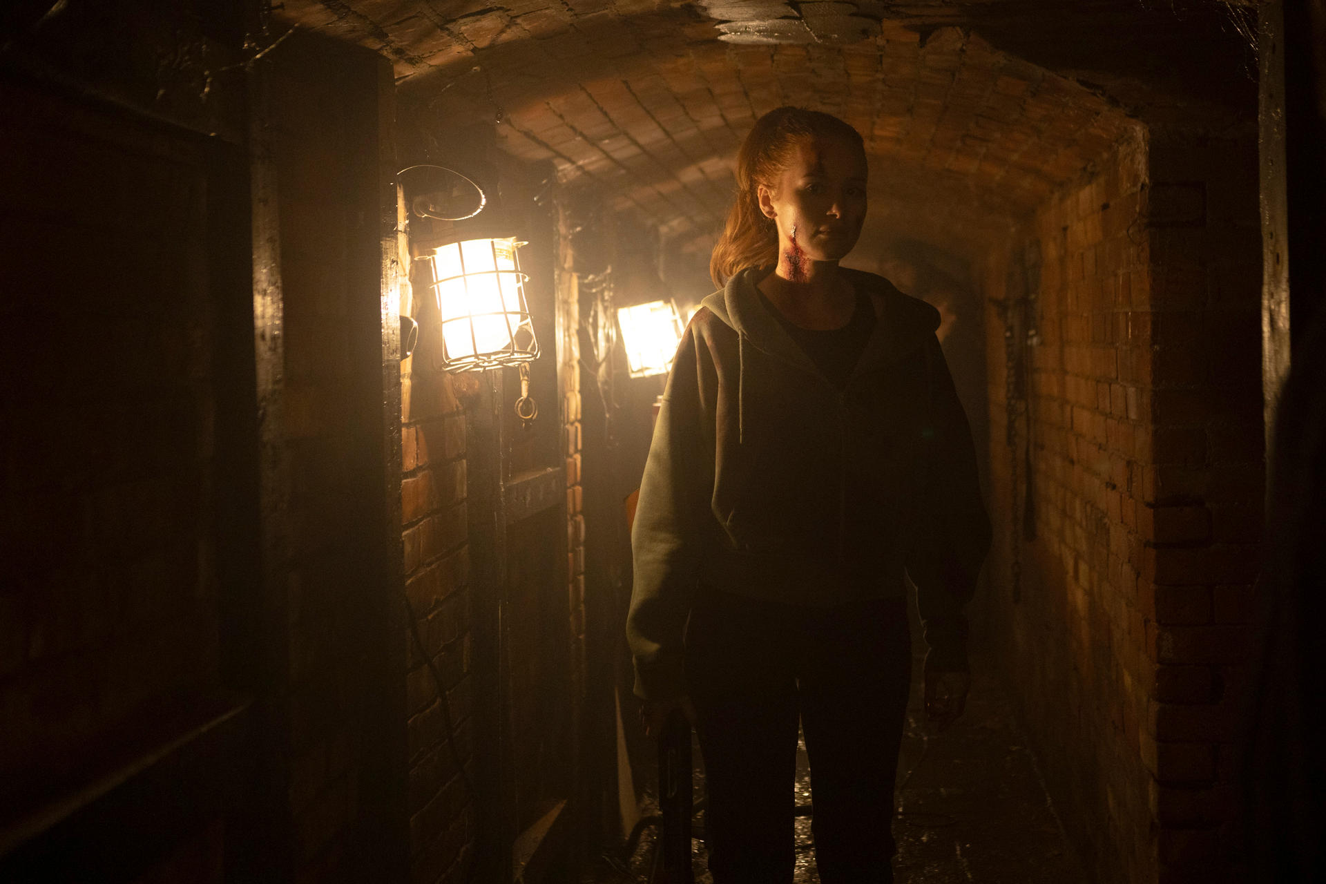 Eine Frau mit roten Striemen am Hals steht in einem dunklen, gemauerten Tunnel.