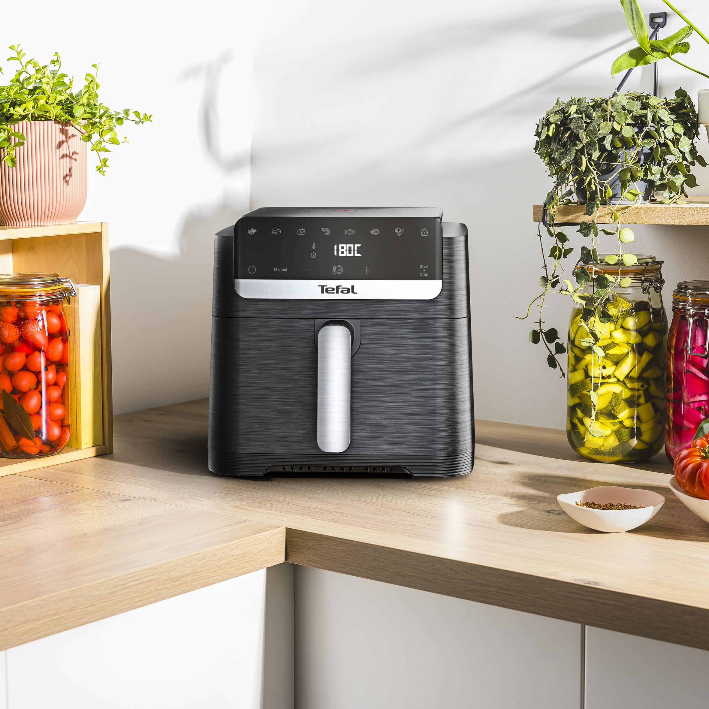 Egy fekete Tefal légkeveréses sütő fa felületen, üvegekkel és növényekkel.
