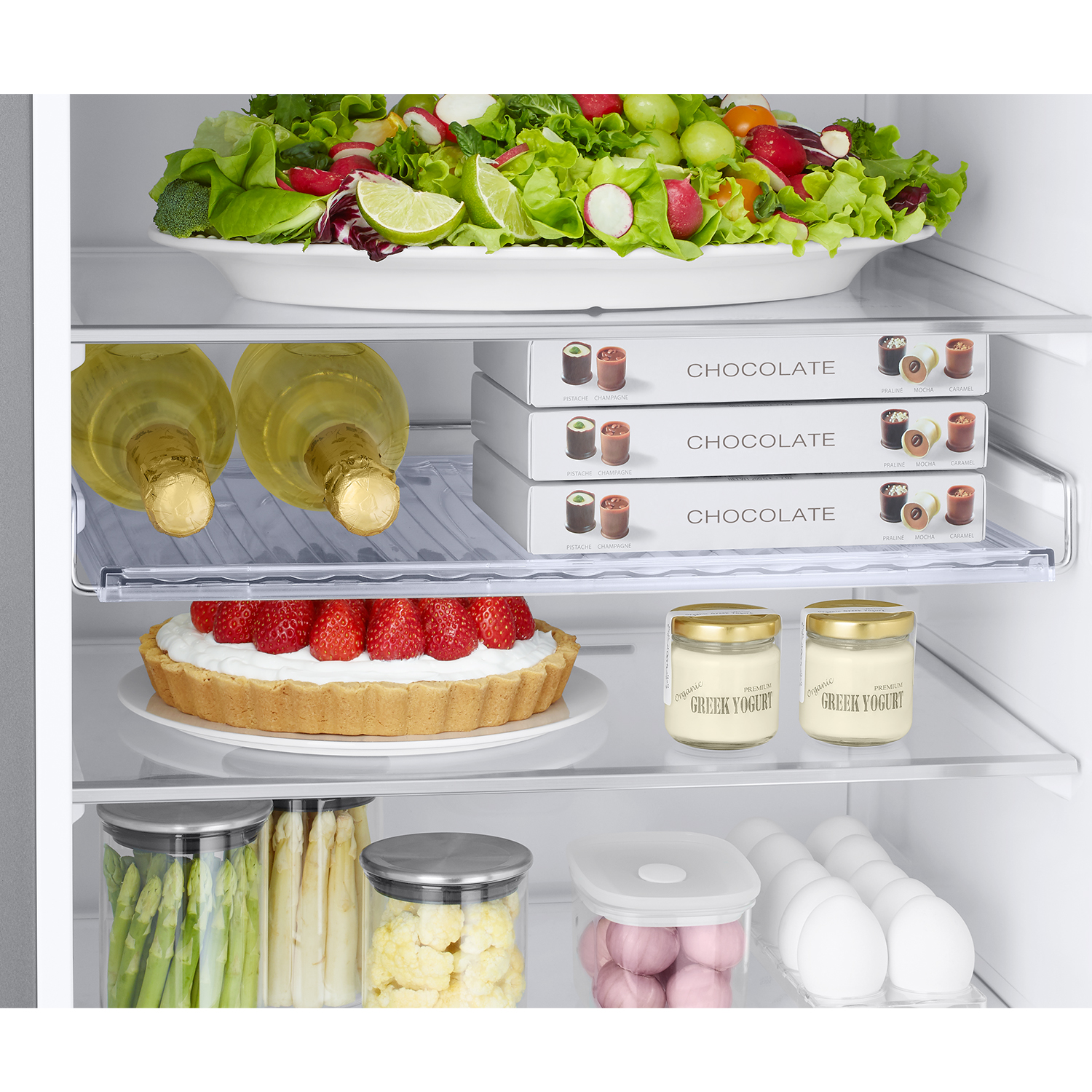 Interior de refrigerador con ensalada, vino, chocolates, tarta, yogur, huevos y verduras.