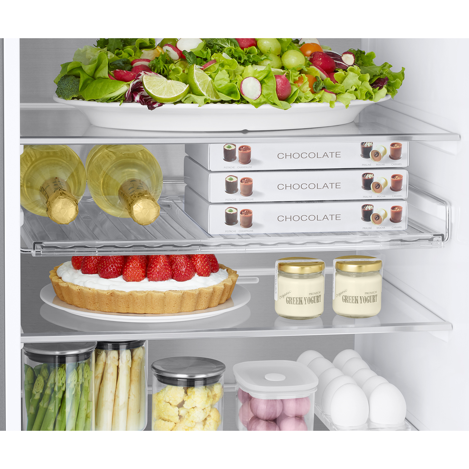 Una repisa de refrigerador tiene ensalada, botellas, chocolates, tarta, yogur, verduras y huevos.