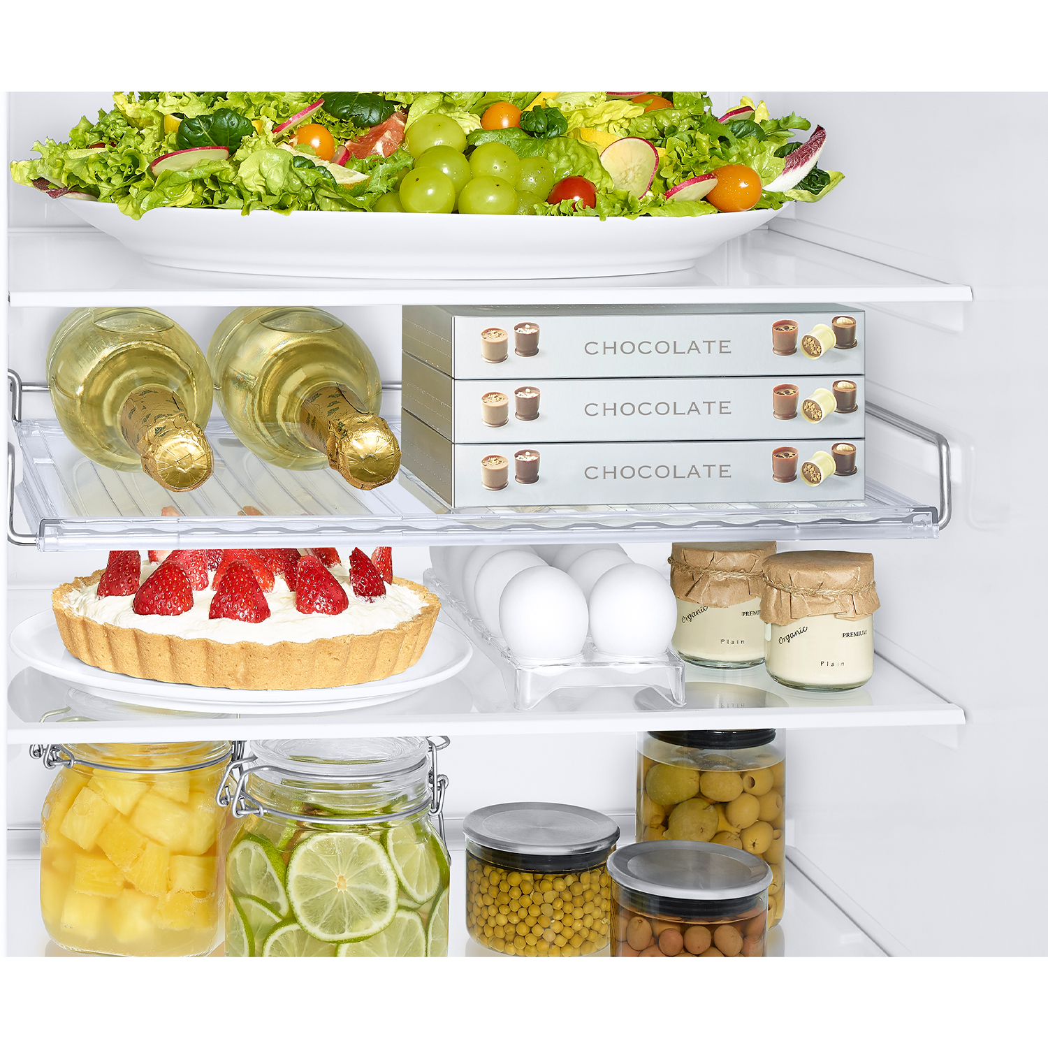 Un estante de refrigerador con ensalada de frutas, chocolates, pastel, huevos y frascos de fruta conservada.
