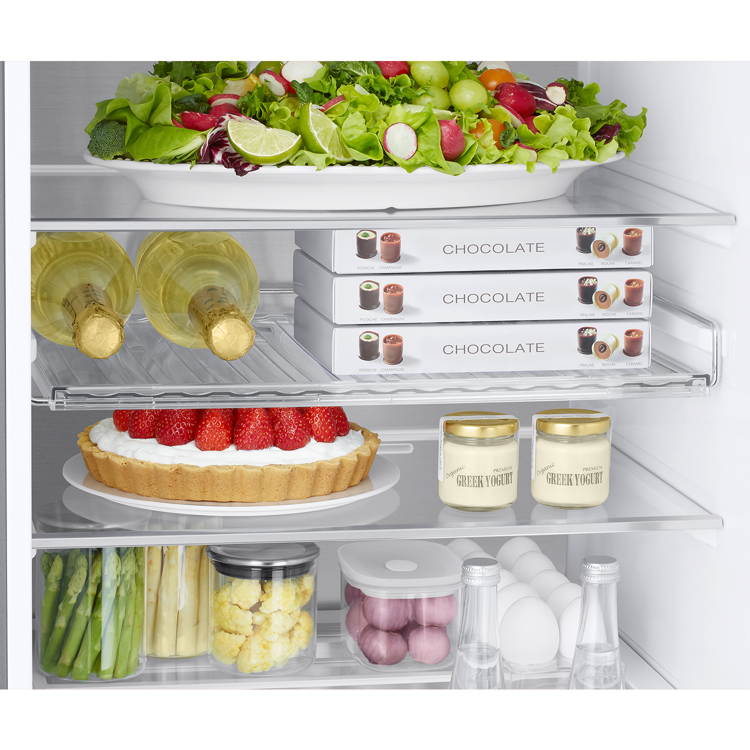 Estantes de refrigerador con ensalada, chocolates, vino, pastel, yogur, verduras y huevos.