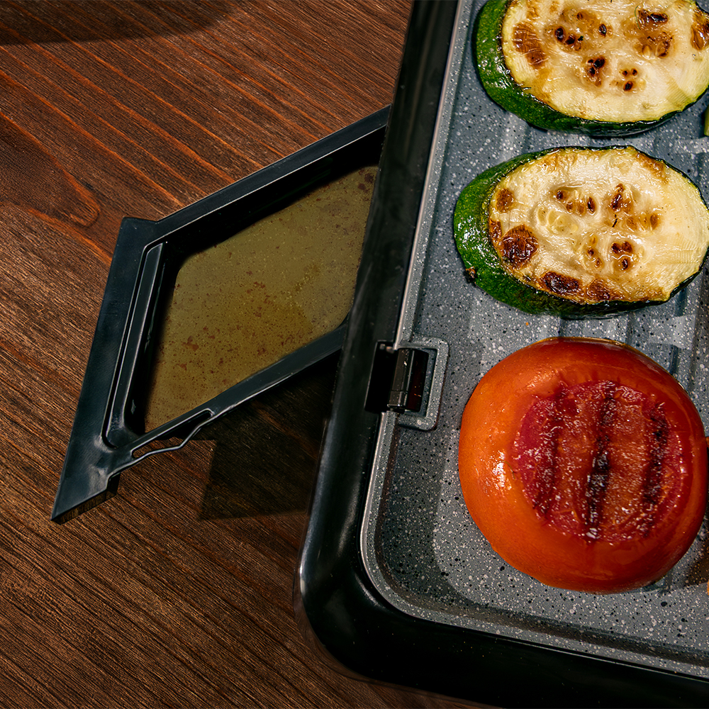Rodajas de calabacín y un tomate a la parrilla en una parrilla eléctrica con bandeja de grasa.