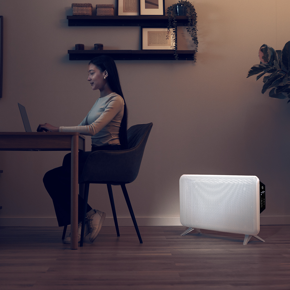 Mujer trabaja en laptop en escritorio, calefactor blanco cerca.
