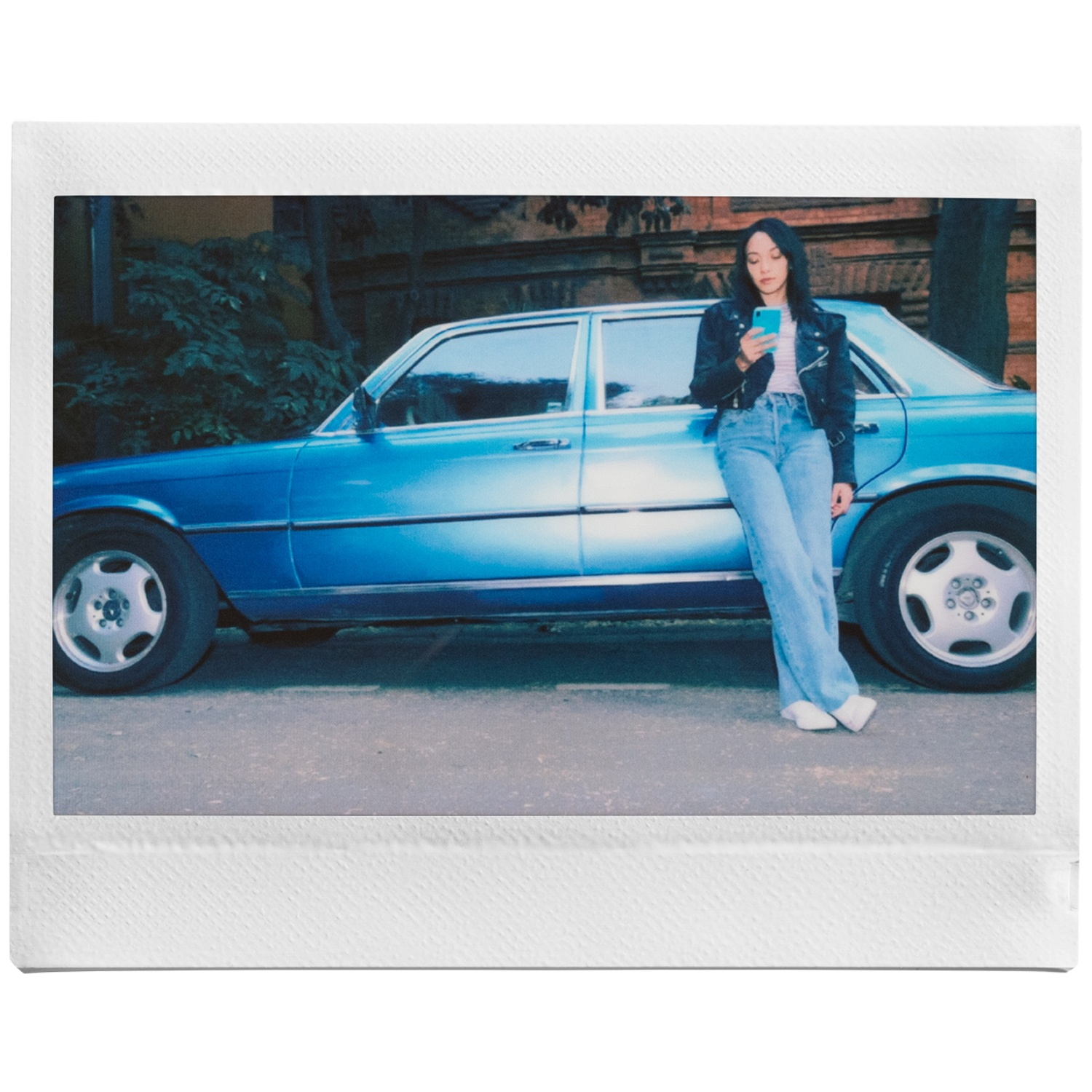 Een vrouw in een spijkerbroek en jas leunt tegen een blauwe auto en kijkt op haar telefoon.