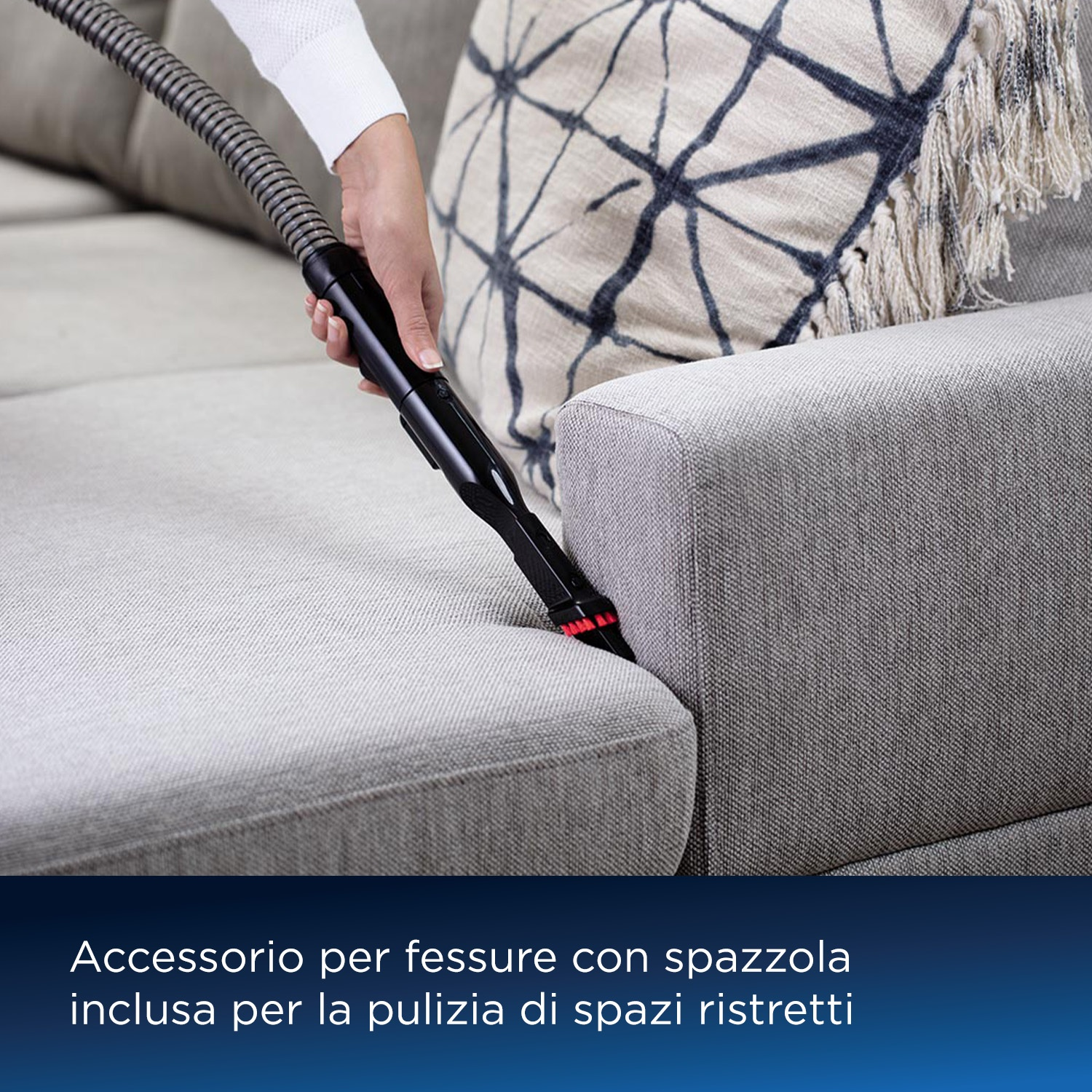 Una mano usa un accessorio per fessure con spazzola per pulire un divano in tessuto grigio.