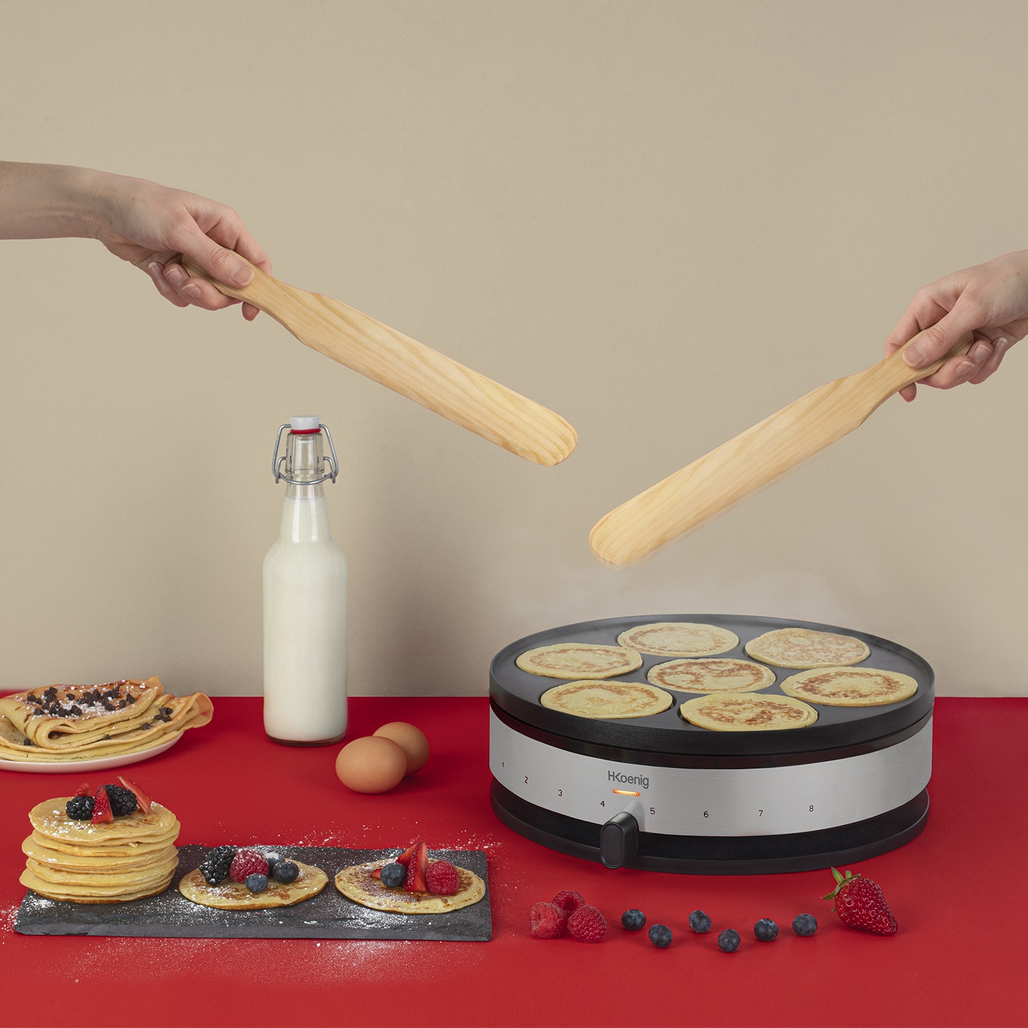 Due mani tengono spatole di legno su una macchina per pancake con pancake, bacche e latte nelle vicinanze.