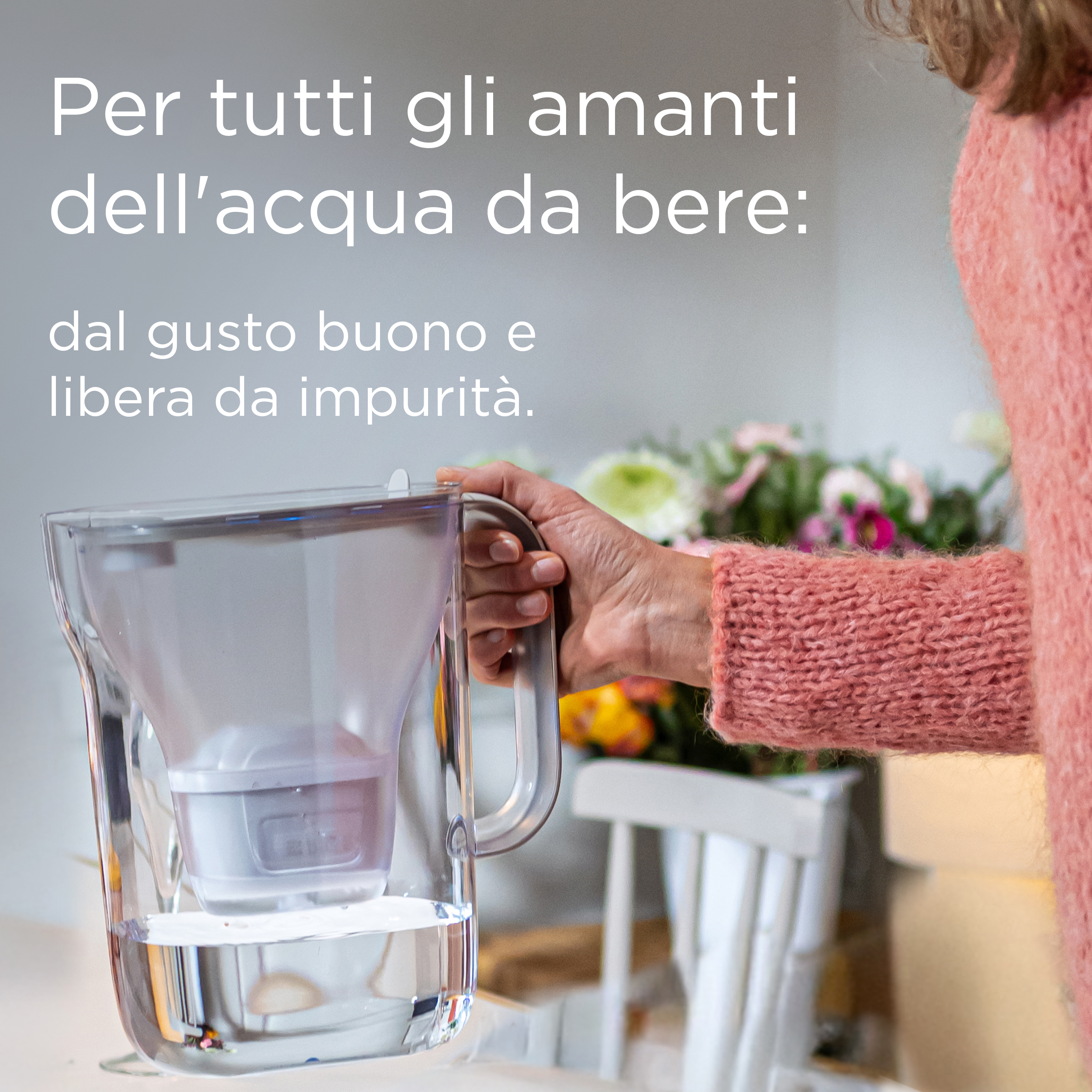 Una mano tiene una brocca filtrante con acqua limpida. Fiori sullo sfondo.