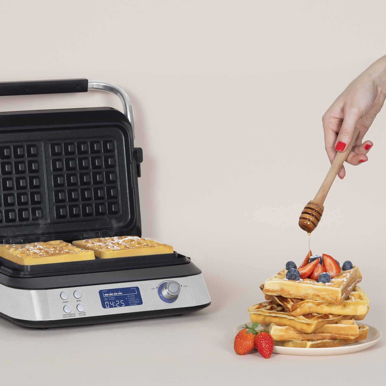 Una piastra per waffle cuoce waffle accanto a una pila di waffle con frutta e miele.