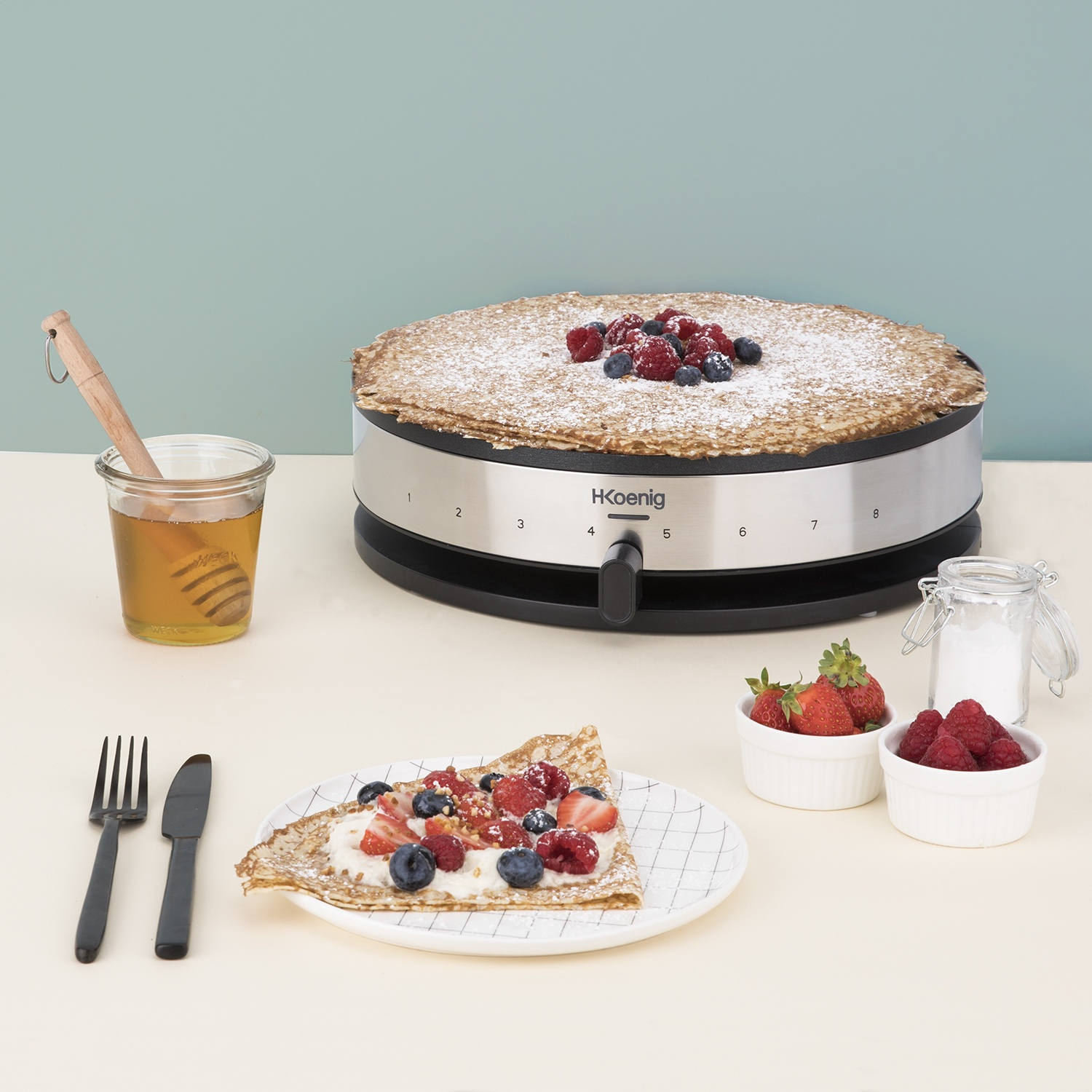 Una grande crêpe cosparsa di zucchero a velo e guarnita con frutti di bosco, vicino a frutta, miele e posate.