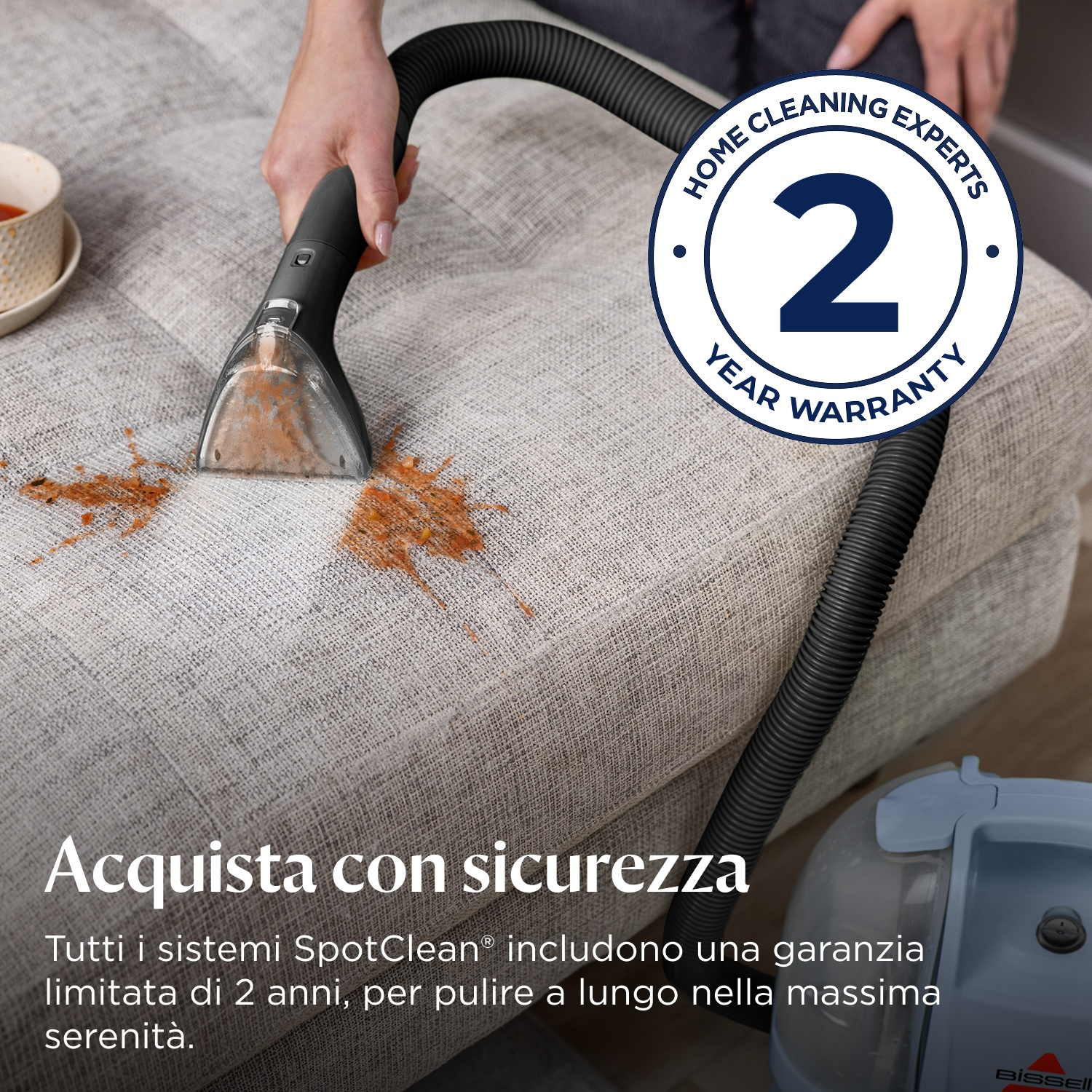 Una mano usa uno strumento per pulire un divano macchiato. Sigillo di garanzia di 2 anni visibile.
