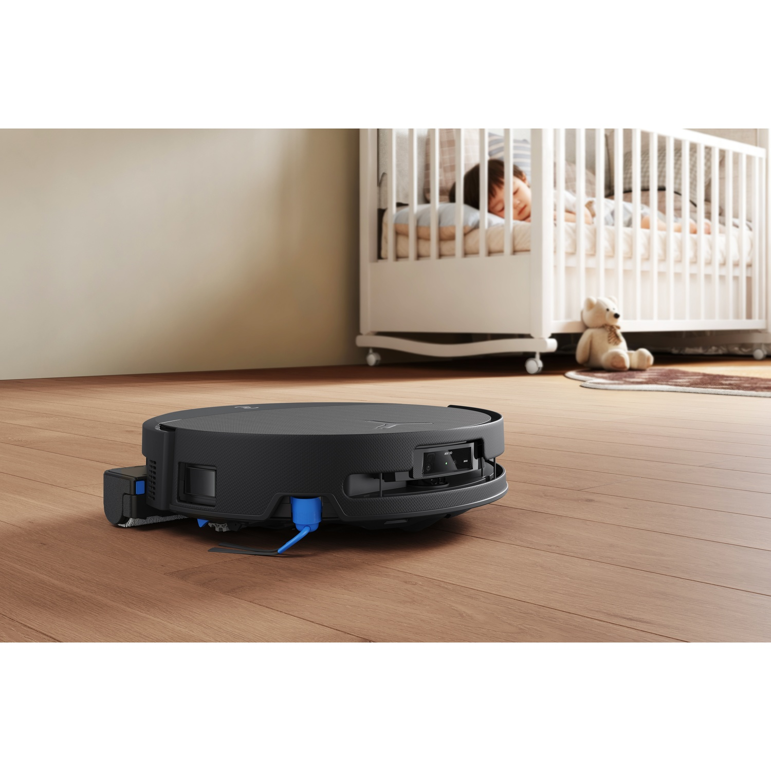 Un robot aspirapolvere nero su un pavimento in legno davanti a una culla dove dorme un bambino.