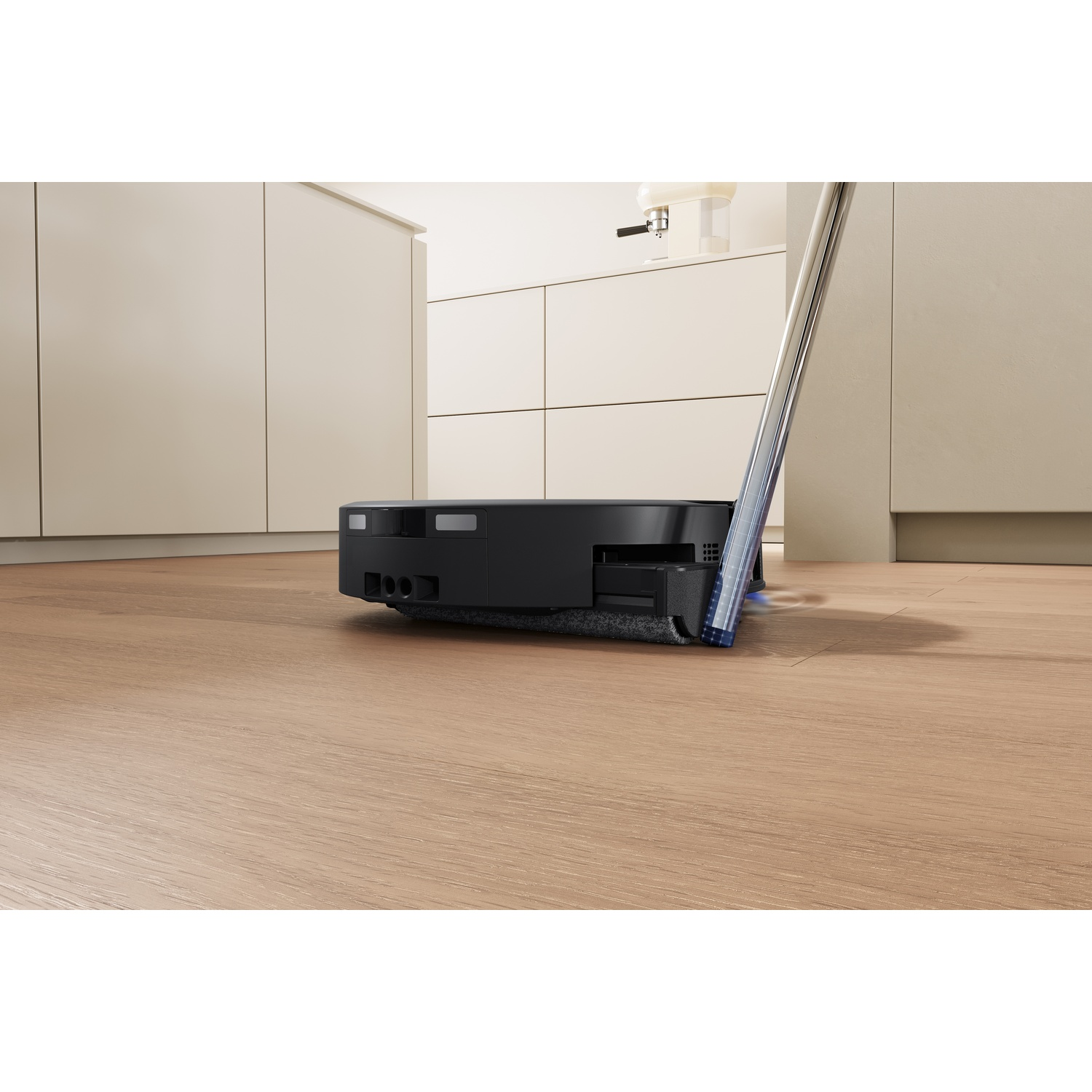 Un robot aspirapolvere nero con funzione lavapavimenti pulisce un pavimento in legno in una cucina moderna.