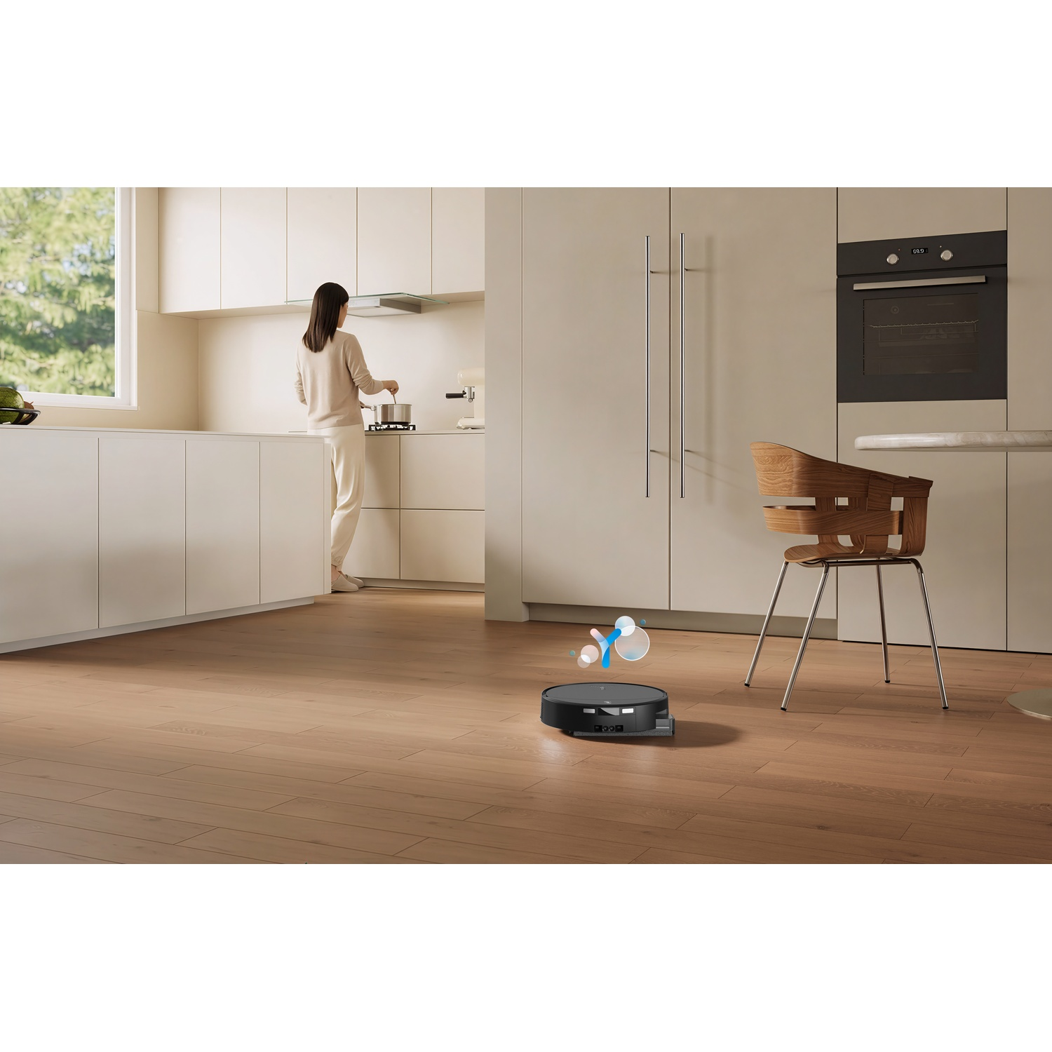 Un robot aspirapolvere pulisce un pavimento in legno in una cucina moderna mentre una donna cucina.