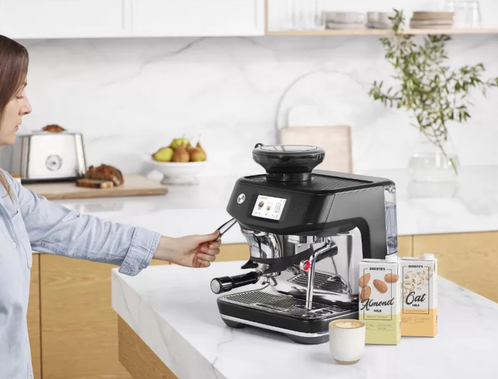 Kobieta obsługuje ekspres do kawy w kuchni. Obok stoją kartony z mlekiem.