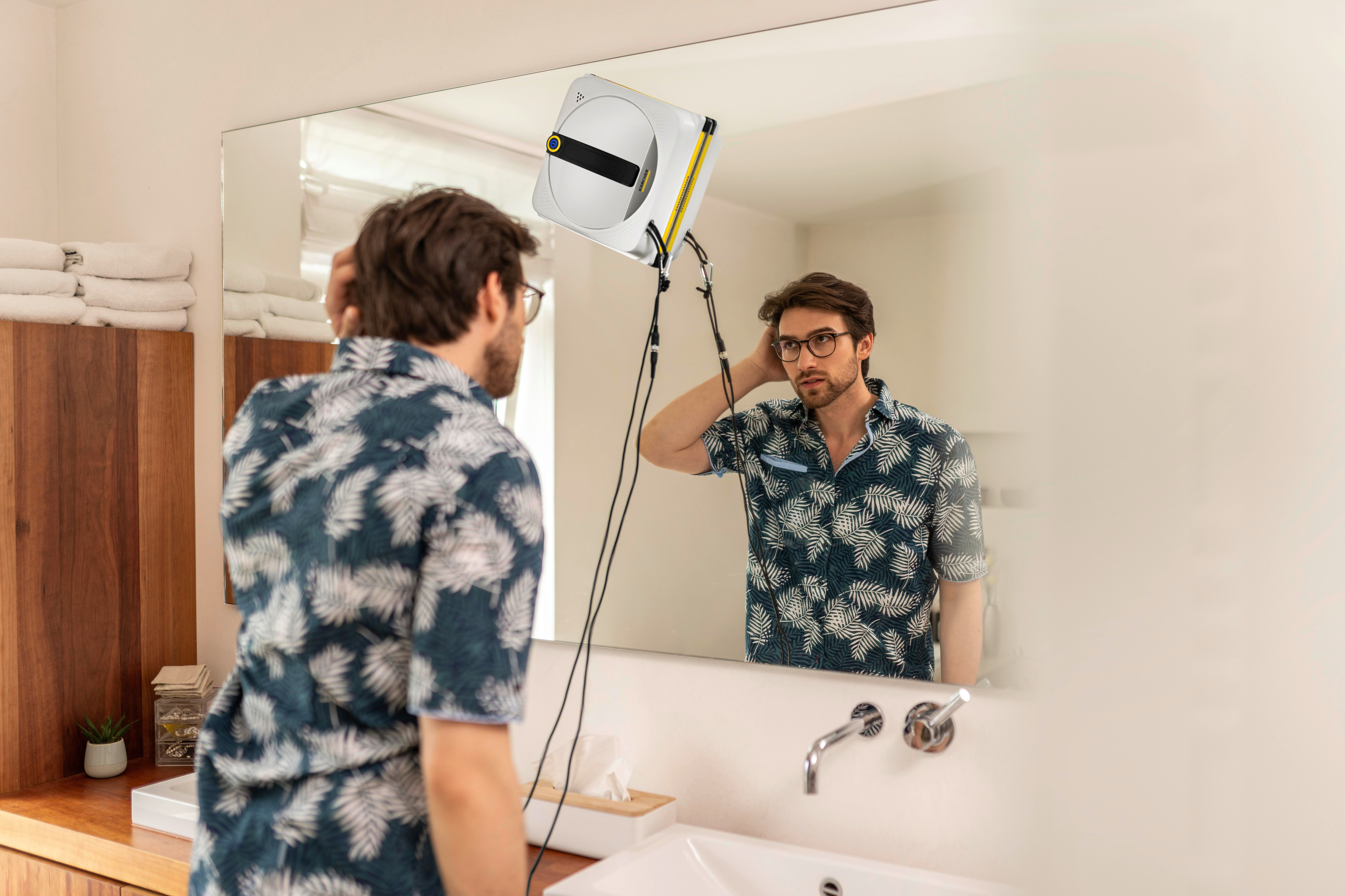 Ein Mann mit Brille blickt in einen Spiegel. Ein Fensterputzroboter hängt daran.