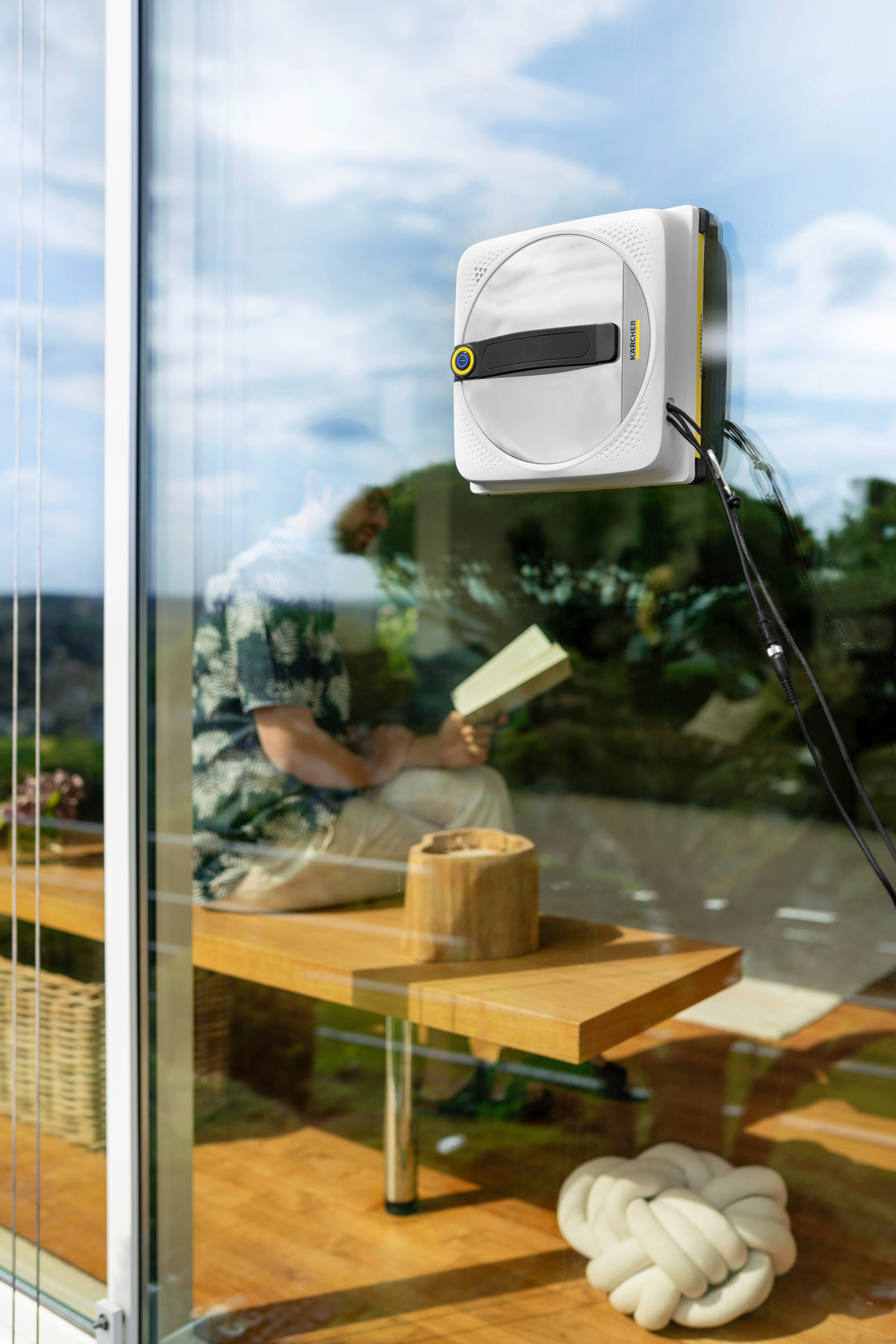 Ein Fensterputzroboter reinigt ein Glasfenster, ein Mann liest im Hintergrund.