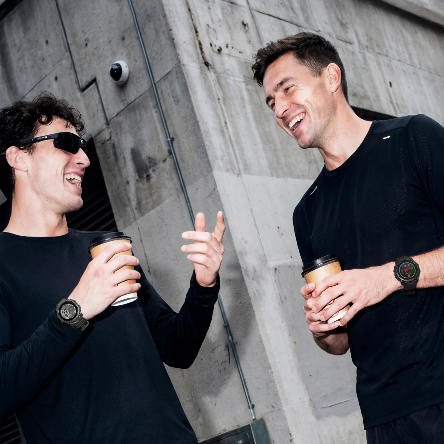 Twee mannen met koffiebekers lachen voor een betonnen muur. Eén draagt een zonnebril.