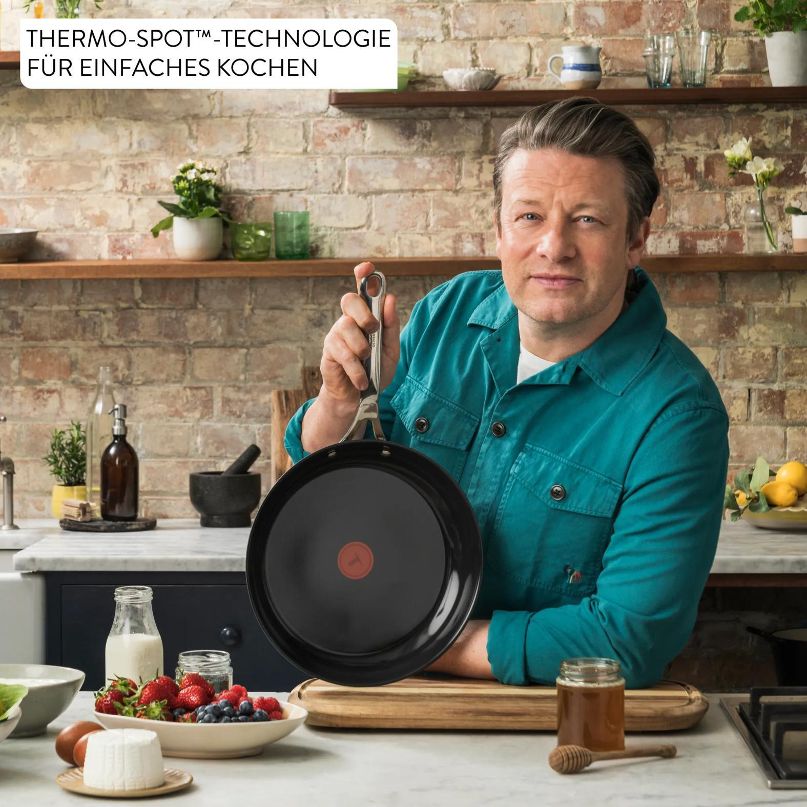 Jamie Oliver hält eine schwarze Pfanne mit rotem Thermo-Spot. Zutaten sind auf dem Tisch.