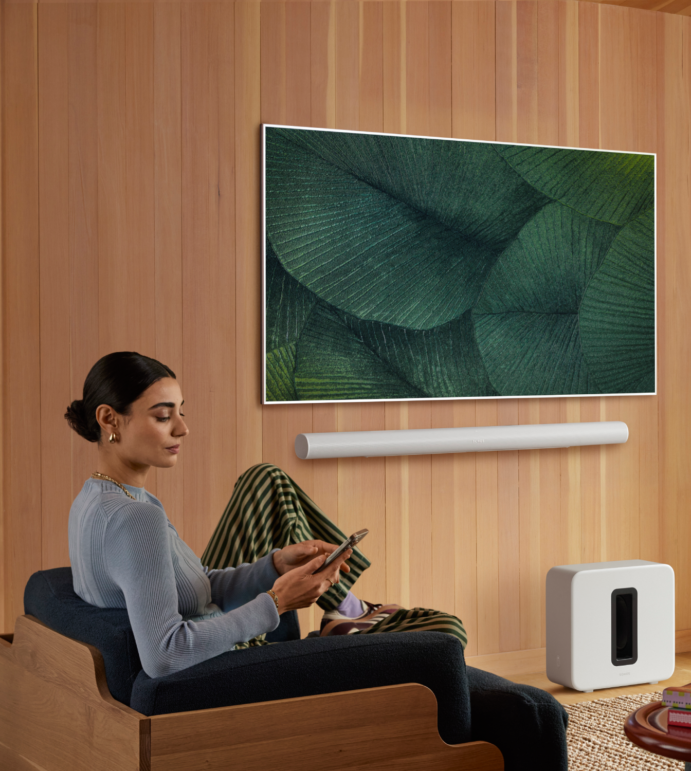 Mujer en sillón usa teléfono, cerca de TV con arte de hojas, barra de sonido y subwoofer.