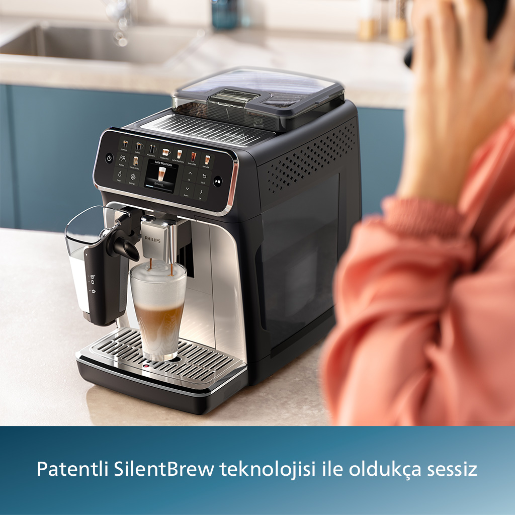 Bir kahve makinesi bardağa latte macchiato yapıyor. Arka planda pembe kazaklı bir kişi.