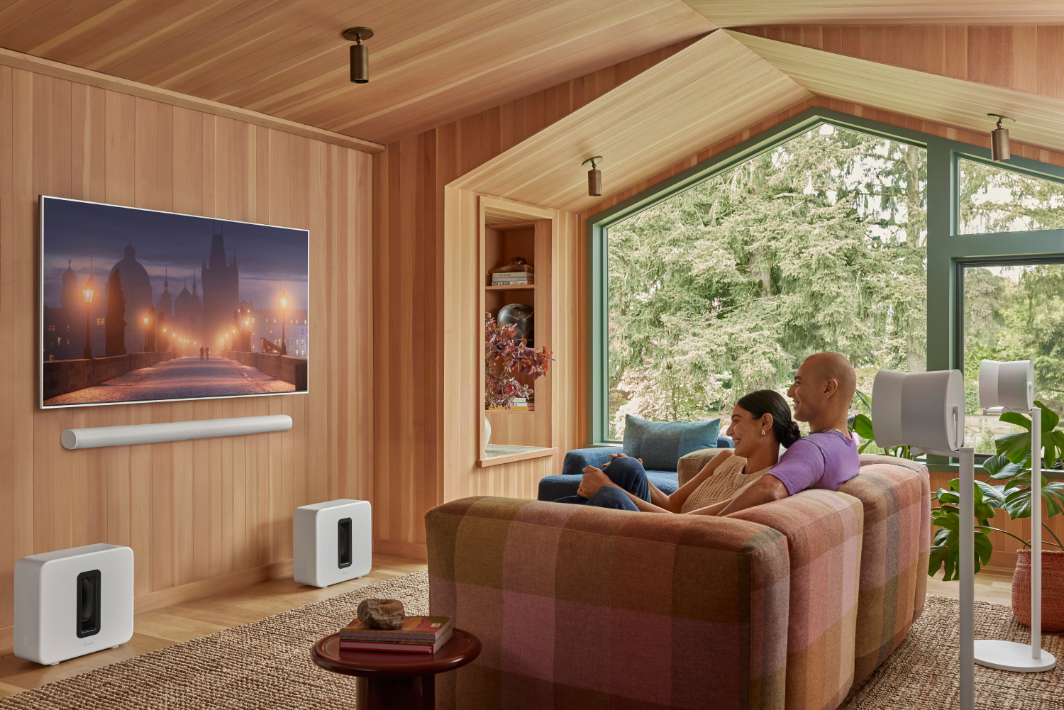 Una pareja se sienta en un sofá en una sala con paneles de madera, TV y altavoces.