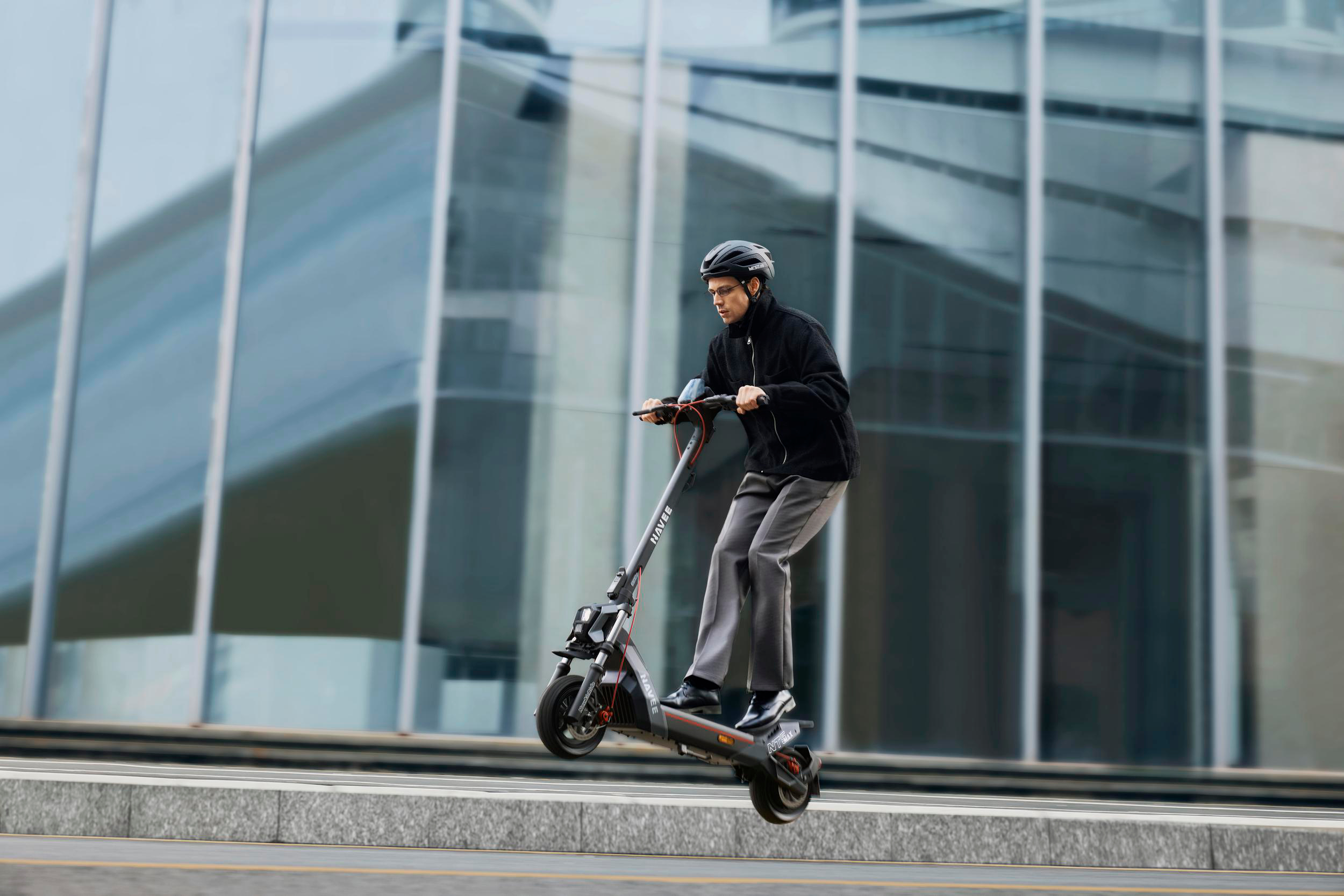 Mann mit Helm fährt E-Scooter vor Glasgebäude.