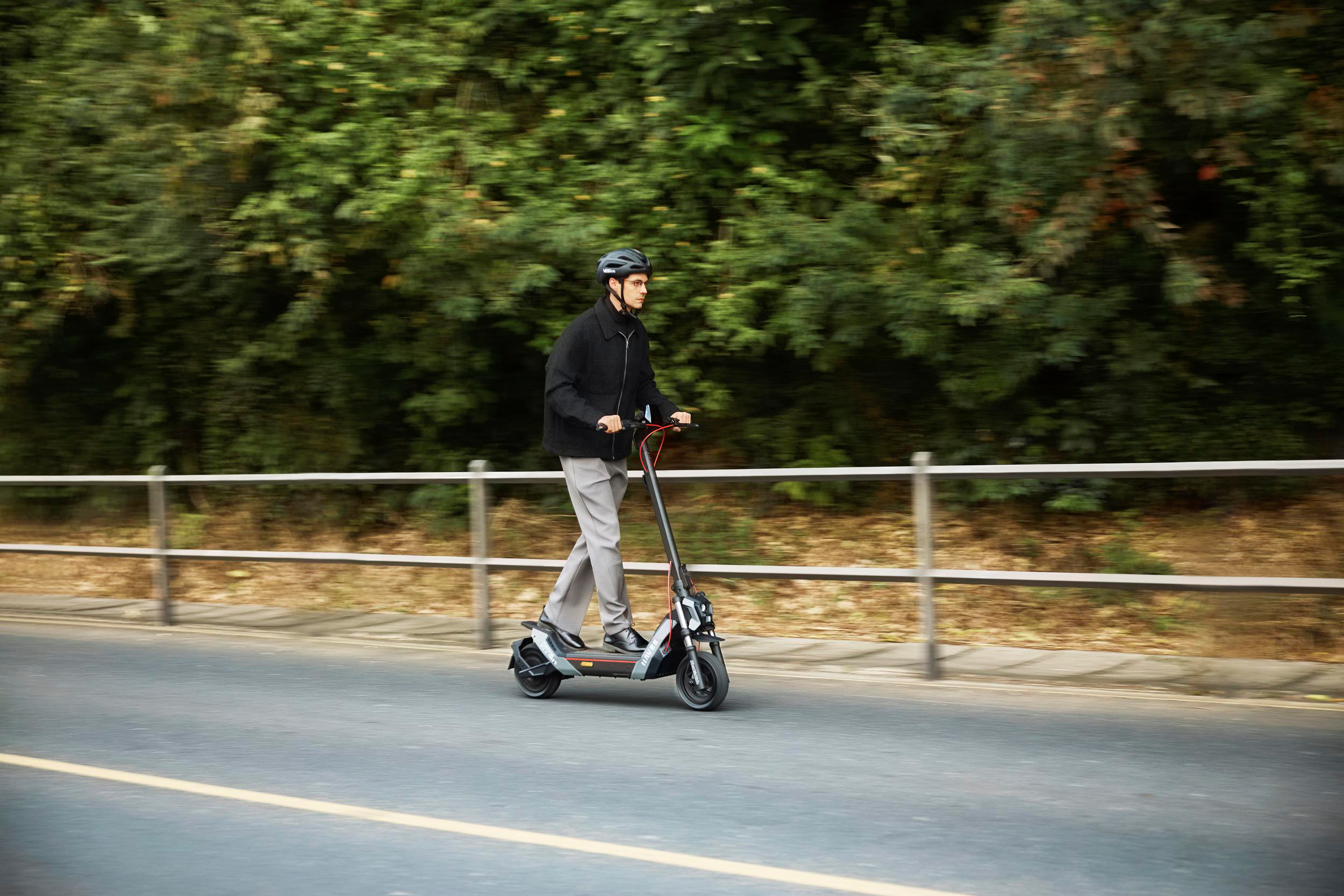 Ein Mann fährt mit einem E-Scooter auf einer Straße, umgeben von Bäumen.