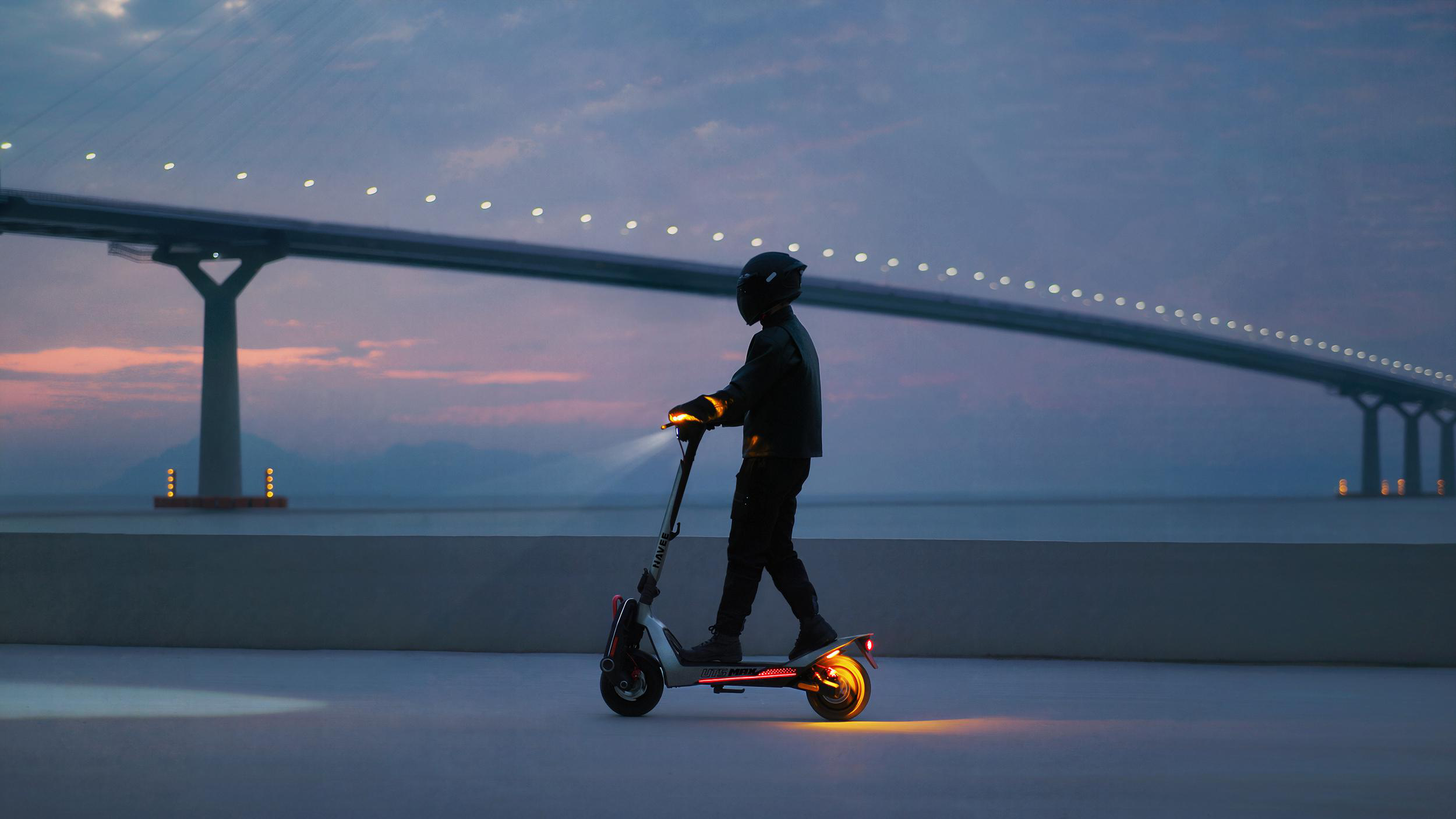 Mann auf E-Scooter fährt bei Dämmerung unter einer beleuchteten Brücke.
