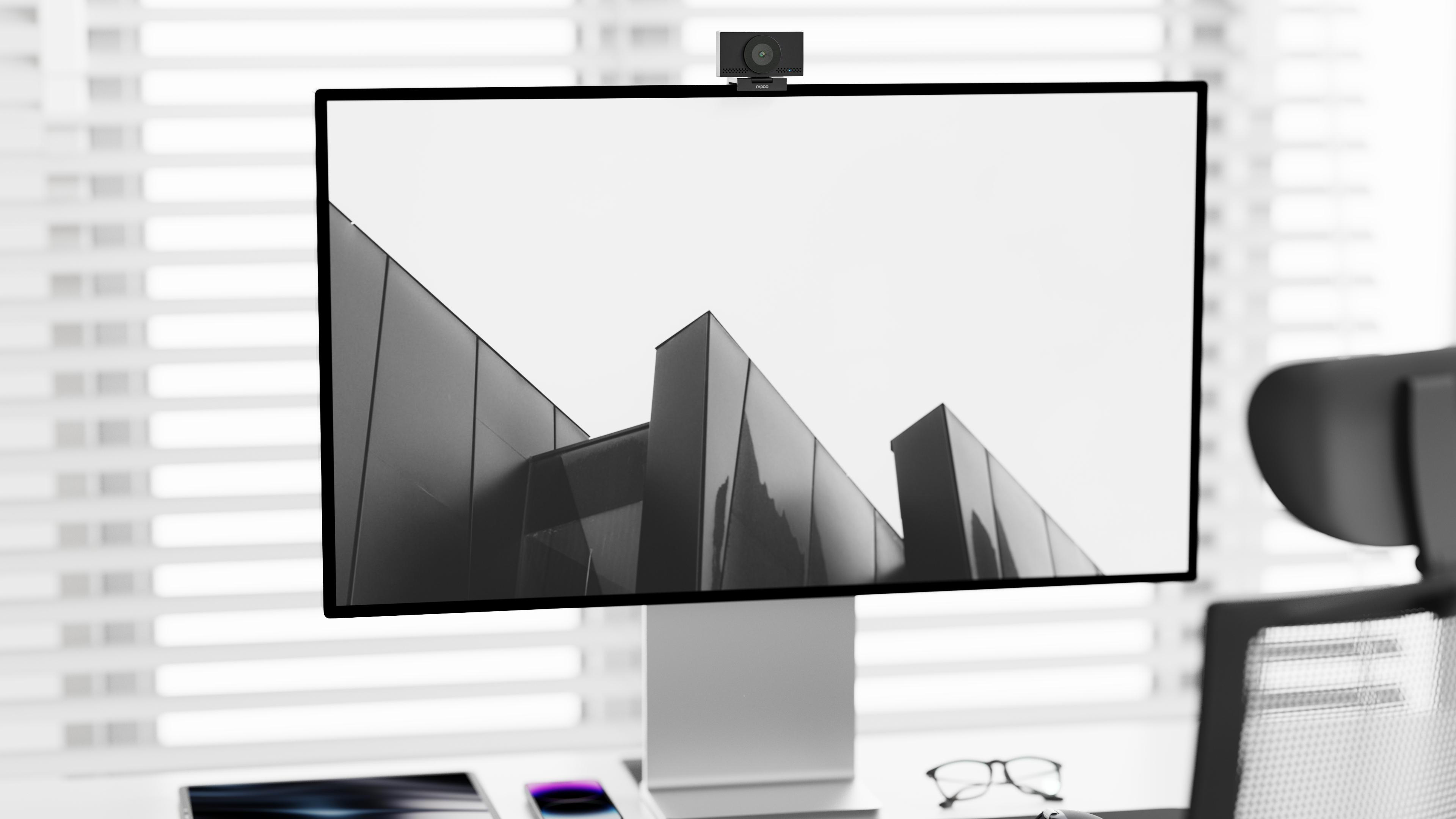 Ein Monitor mit Webcam zeigt eine moderne Architekturansicht. Daneben liegen Brille und Smartphone.