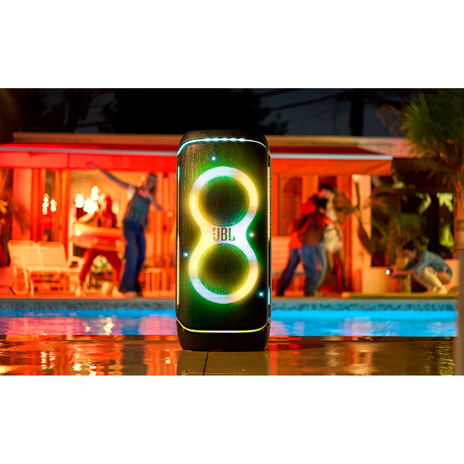 A JBL speaker with light effects sits by a pool at night. People dance in the background.
