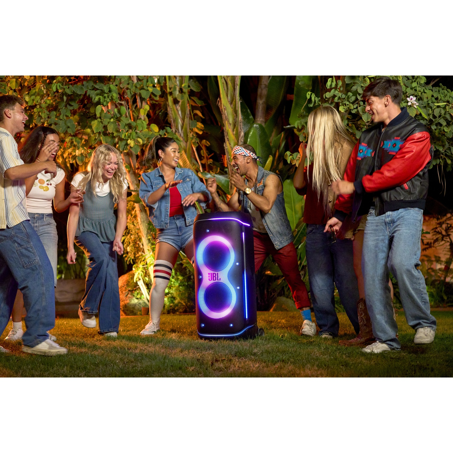 Young people dance outdoors around a JBL speaker with colorful lights.